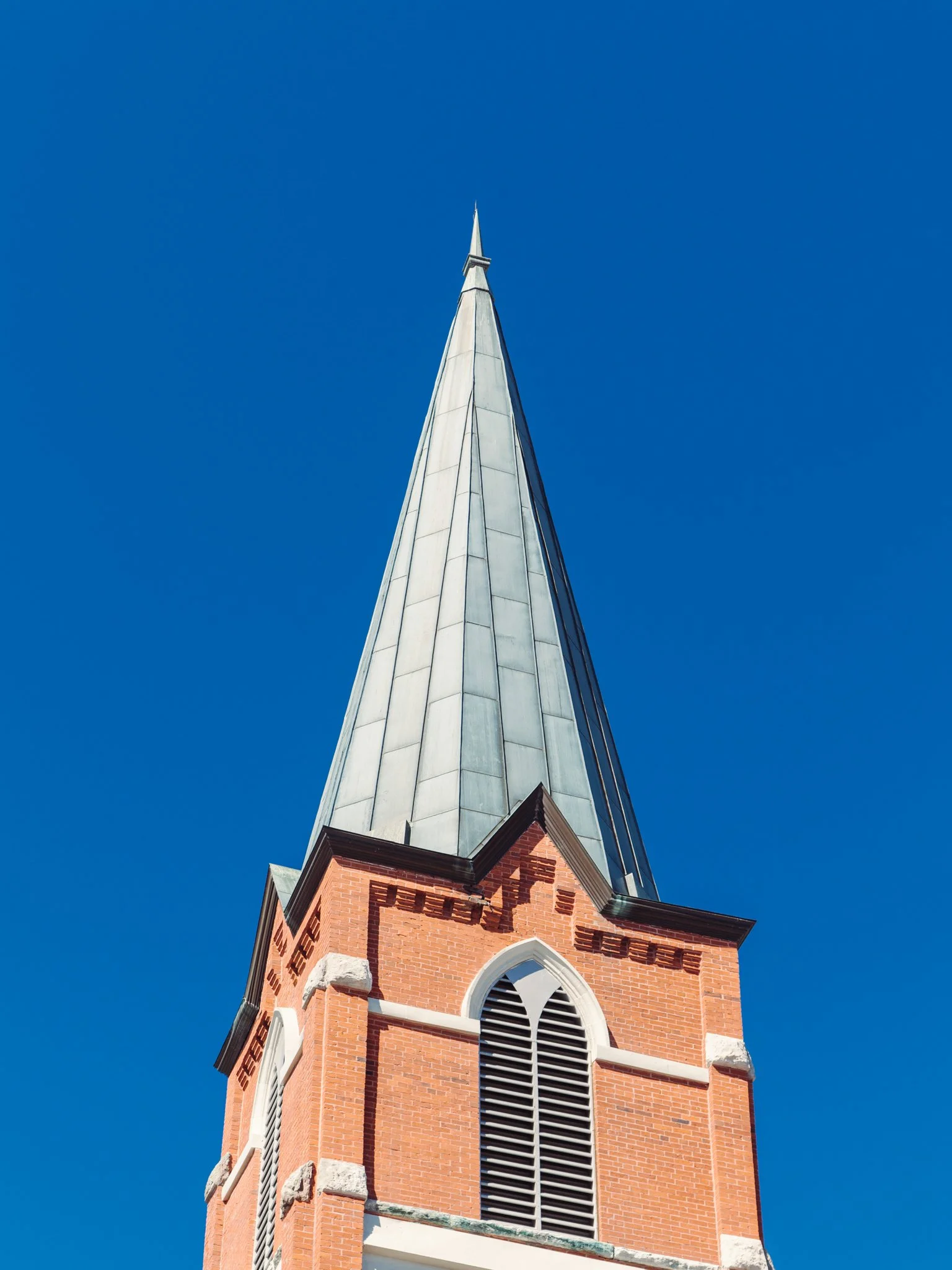 A church steeple with a pointed, metallic roof against a clear blue sky. The church is made of red bricks with arched windows and white trim.