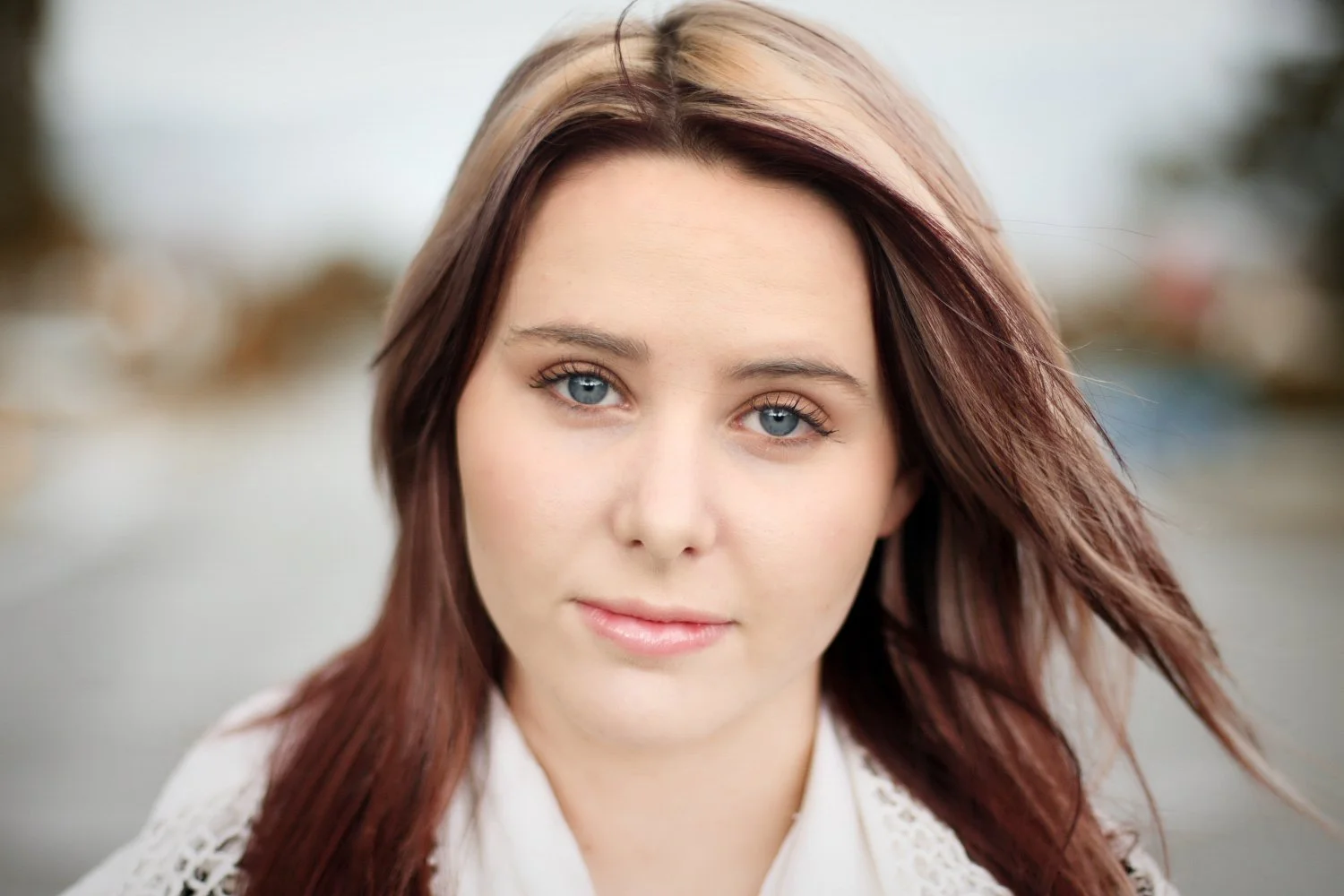 Close-up of a young woman with blue eyes and shoulder-length reddish-brown hair, outdoors with blurred background.