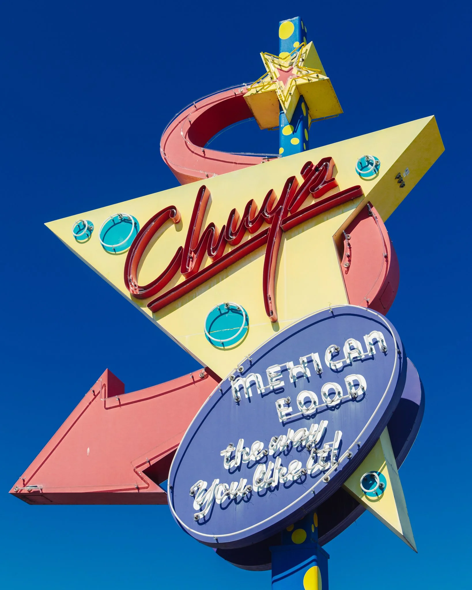 Colorful vintage neon sign against a clear blue sky, featuring the words 'Gambling', 'Games', and 'Eat' with decorative elements including stars and circles.