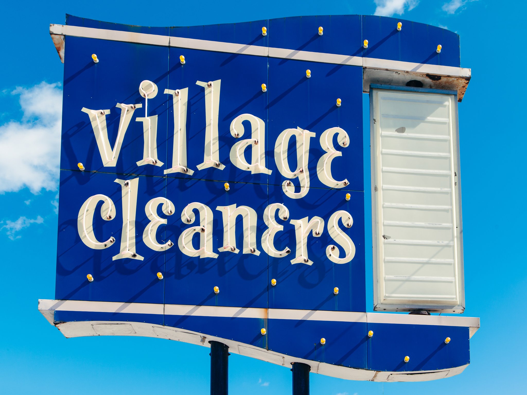 Close-up of a vintage blue and white neon sign with the words "Valley Oasis" against a bright blue sky with some clouds.
