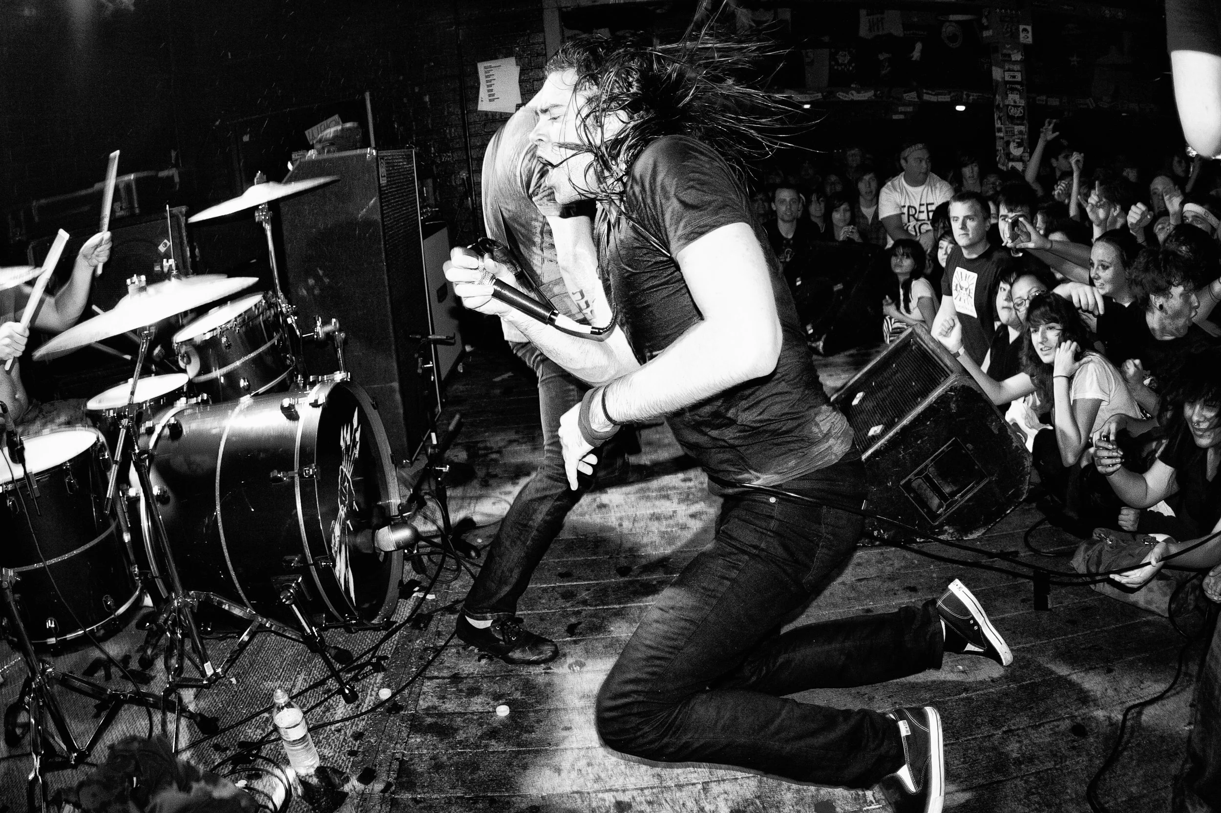 A woman passionately singing into a microphone during a live music concert, with her hair flying. She is kneeling on the stage near a drum set, surrounded by an engaged audience.