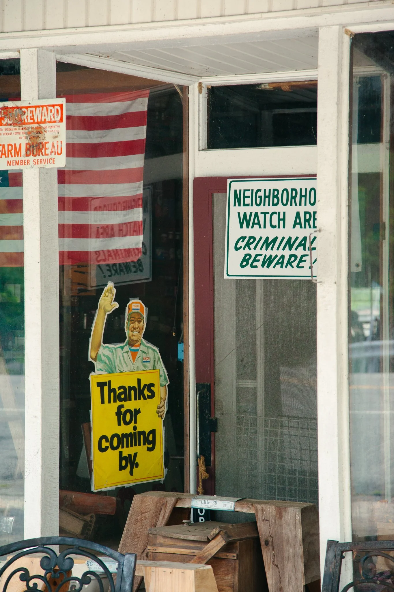 View inside a store or cafe with signs on the door, including a yellow sign saying 'Thanks for coming by,' and a green and white sign that states 'Neighborho watch are criminal beware.' There's a cardboard box or wooden crate on the floor and a decor