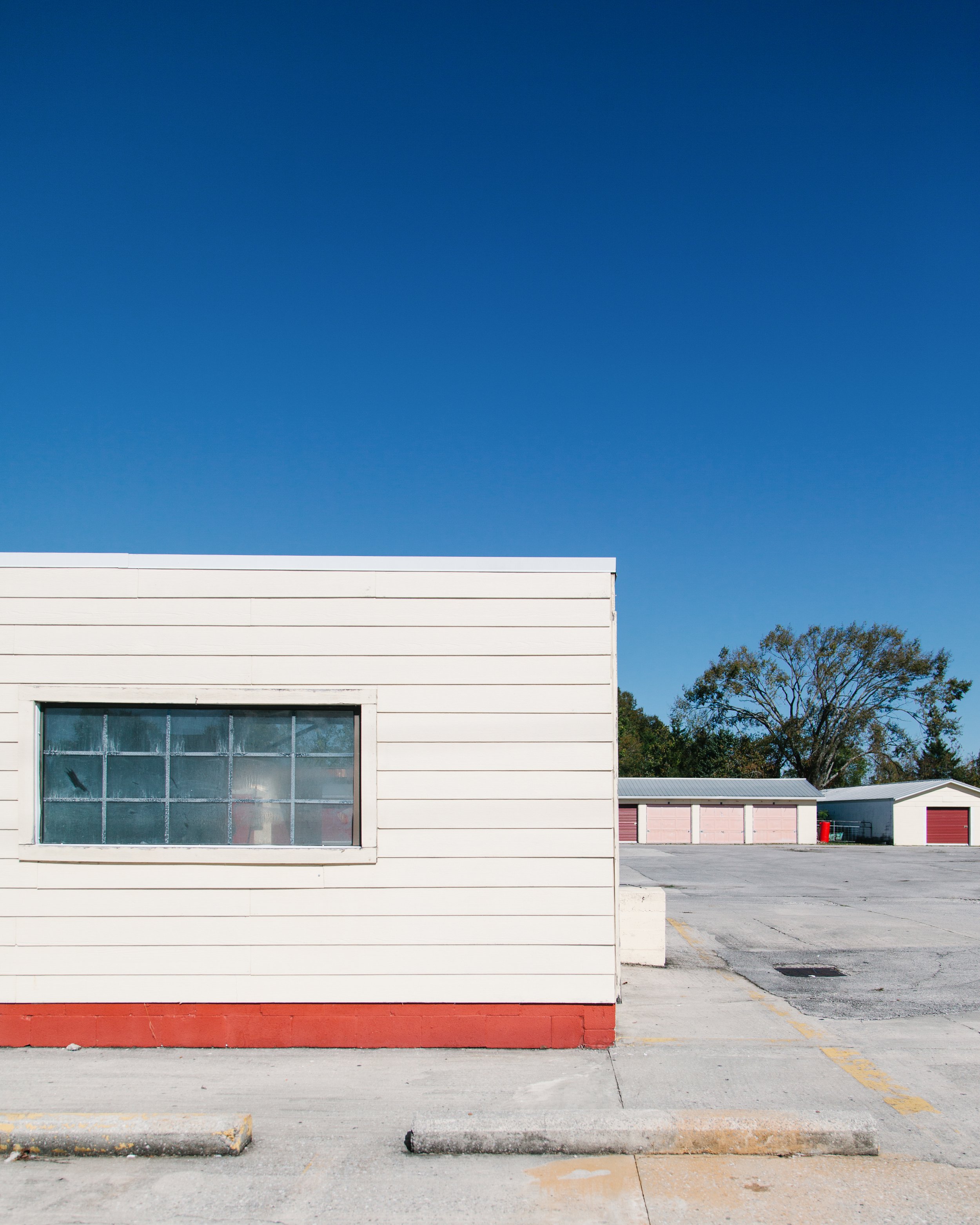 An empty parking lot in front of a white building with a large window and three storage units with pink doors in the background. Clear blue sky overhead and some trees on the right side.