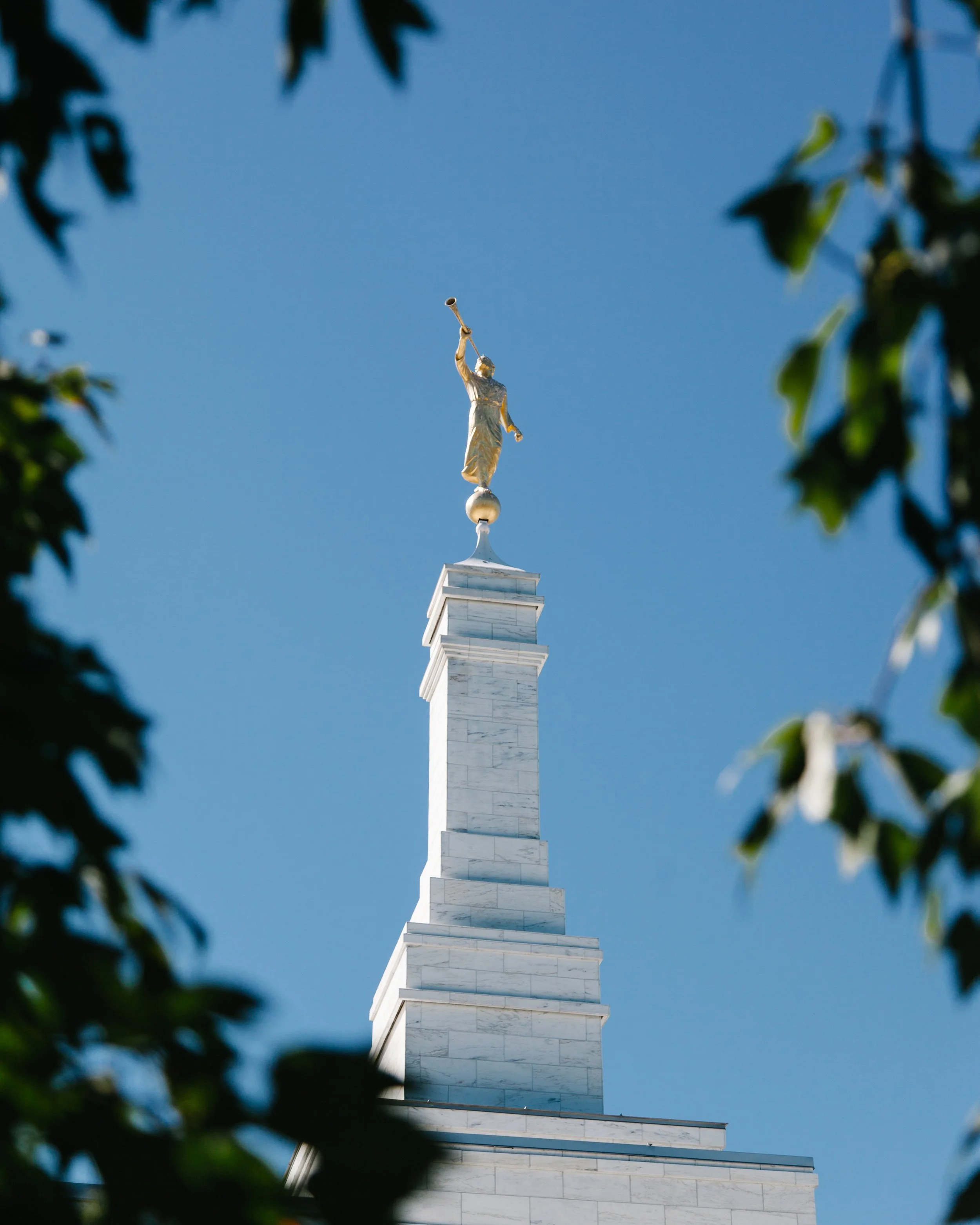 Statue of a woman holding a trumpet, atop a tall marble column against a clear blue sky, framed by blurred leaves.