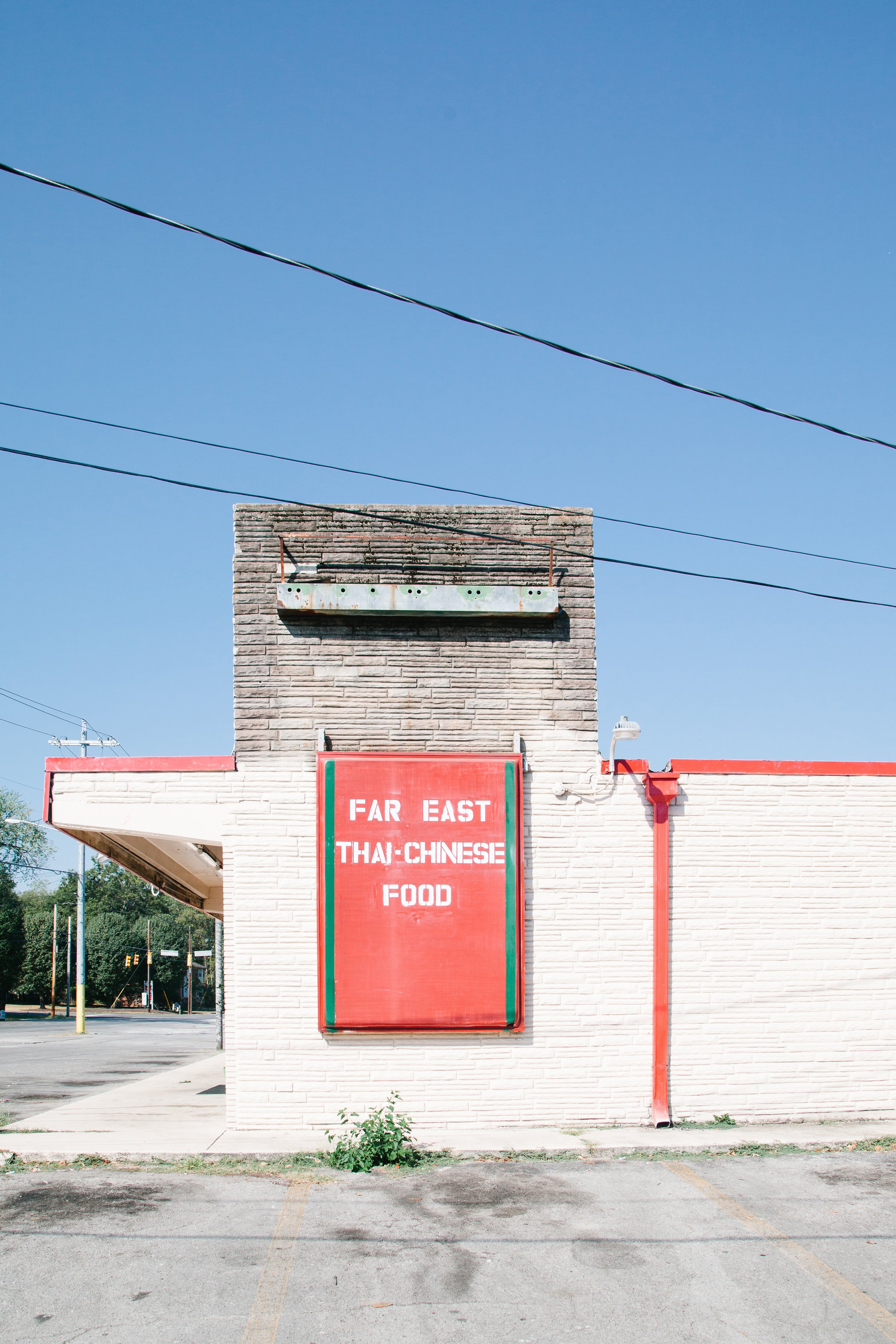 A white brick building with a red and green sign that reads 'FAR EAST THAI-CHINESE FOOD'. The building has a chimney with a metal overhang at the top and electrical wires above it. The surrounding area is a parking lot with marked parking spaces and 