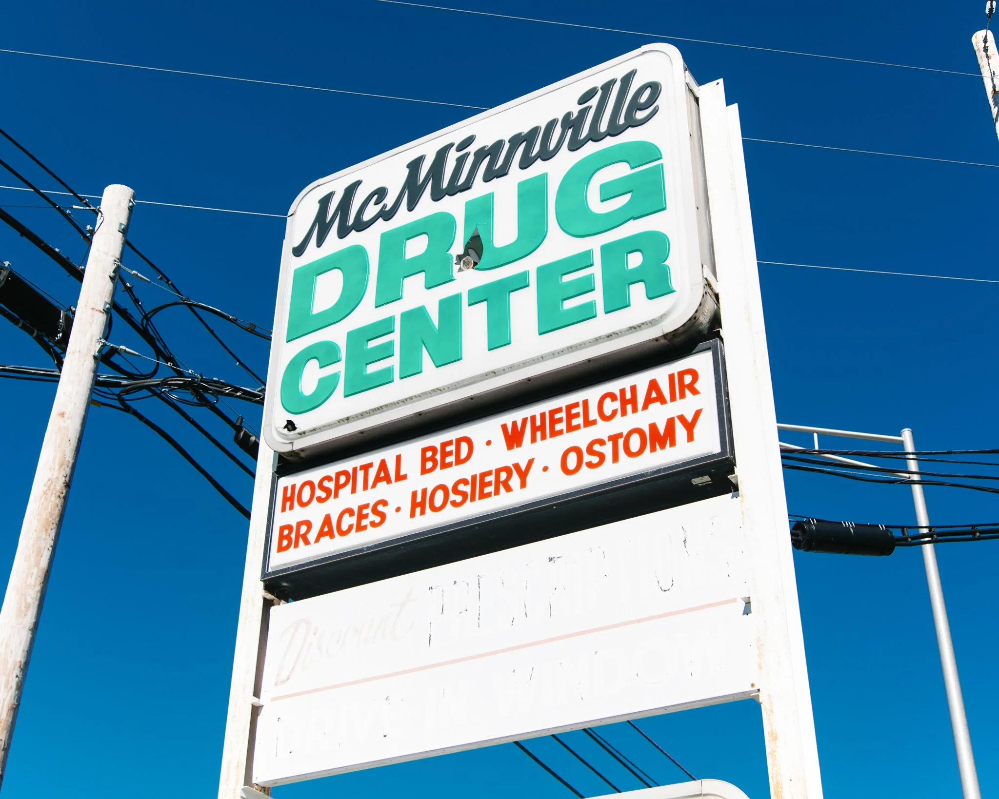 A white sign with green and red text advertising medical and hospital services, mounted on a white pole against a clear blue sky. The sign mentions services such as hospital bed, wheelchair, braces, and ostomy supplies.