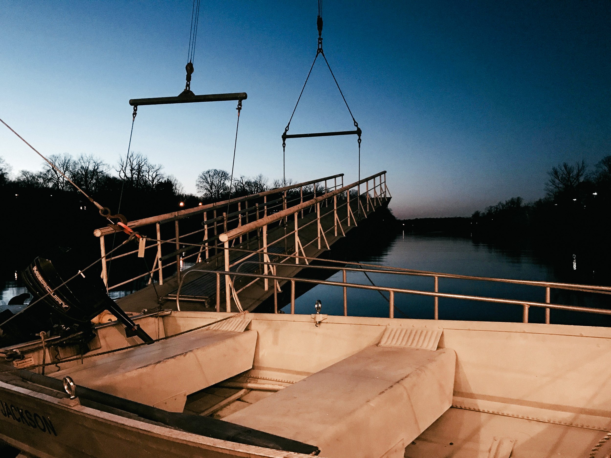 A wooden boat docked at a pier on a calm body of water during dusk, with a boat motor visible at the stern and a metal walkway leading to the shore. Trees are silhouetted in the background against the darkening sky.