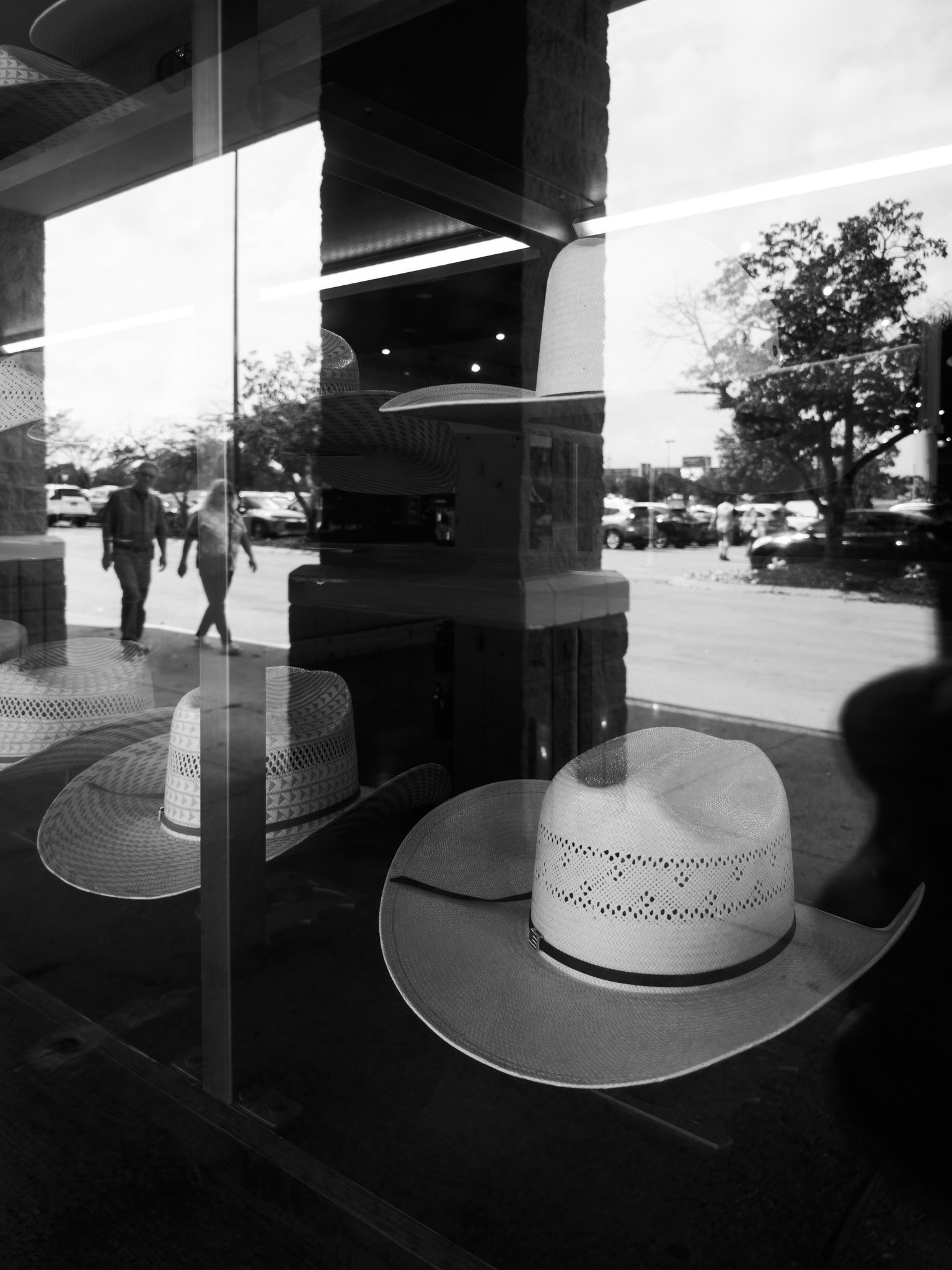 Black and white photo showing the reflection of a store window with woven sun hats and city street outside, trees, cars, and pedestrians.