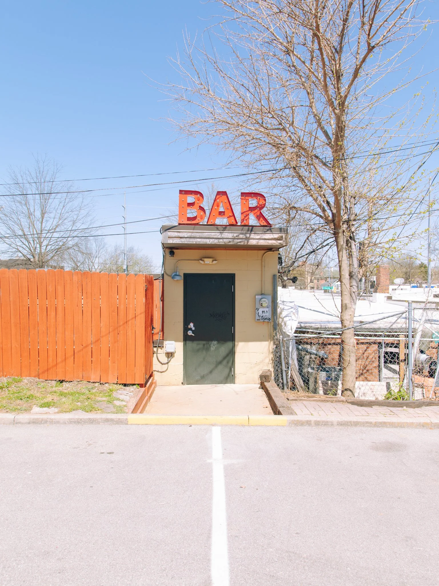 A small building with a sign that reads "BAR" in red lights on top. The building has a black door with some graffiti, a small light, and an electrical box outside. There is a tall tree with few leaves behind the building and a chain-link fence to the