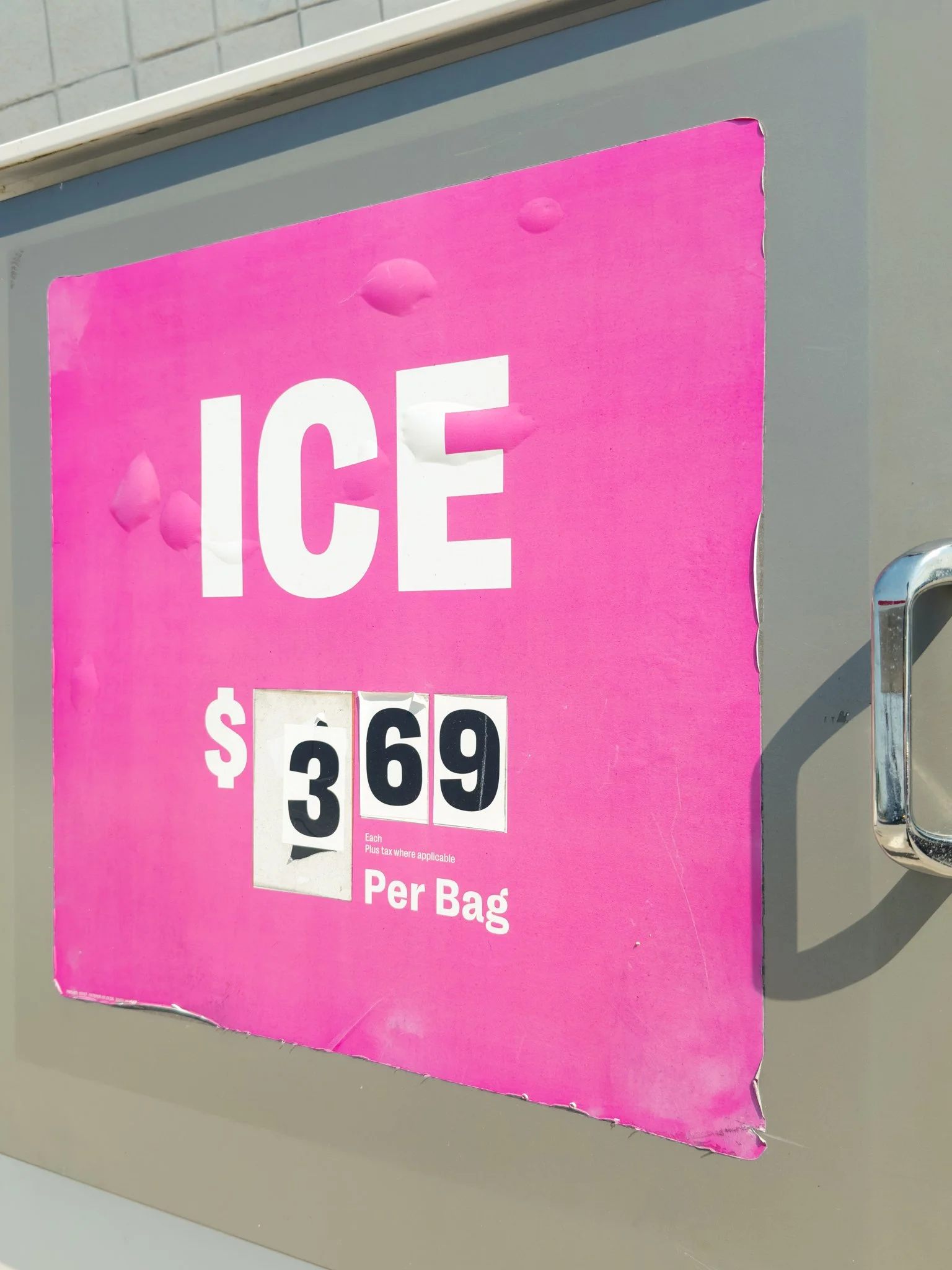 Pink sign indicating ice for $3.69 per bag, with visible damage and peeling near the edges, mounted on a gray surface with a metal handle or pointer visible on the right side.
