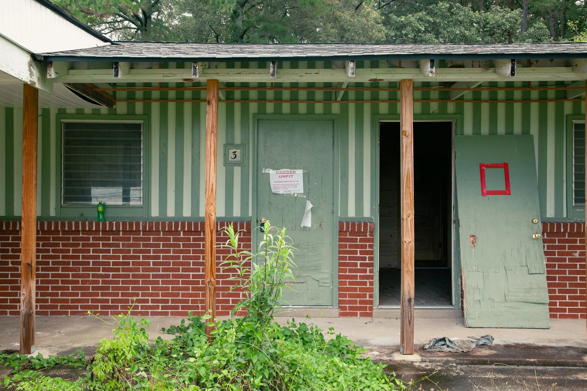Front of a house under renovation, with a partially installed front porch. The house has green siding with vertical stripes, a red brick foundation, and a green door. There are two windows with metal bars. Vertical wooden support beams are in place f