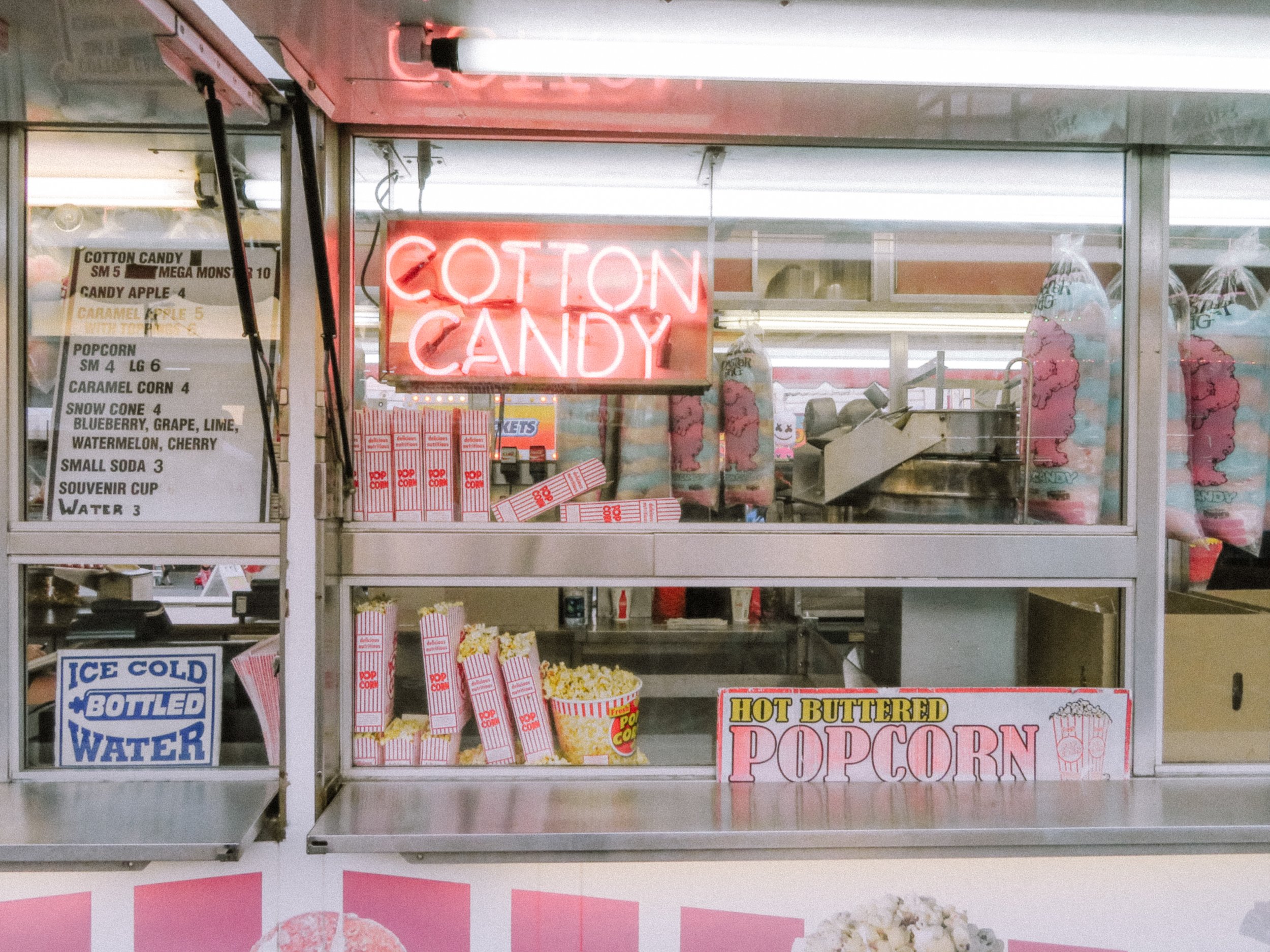 A popcorn stand with a bright pink neon sign that says 'Cotton Candy,' plastic bags of cotton candy on display, and a menu listing various candy and snack options.