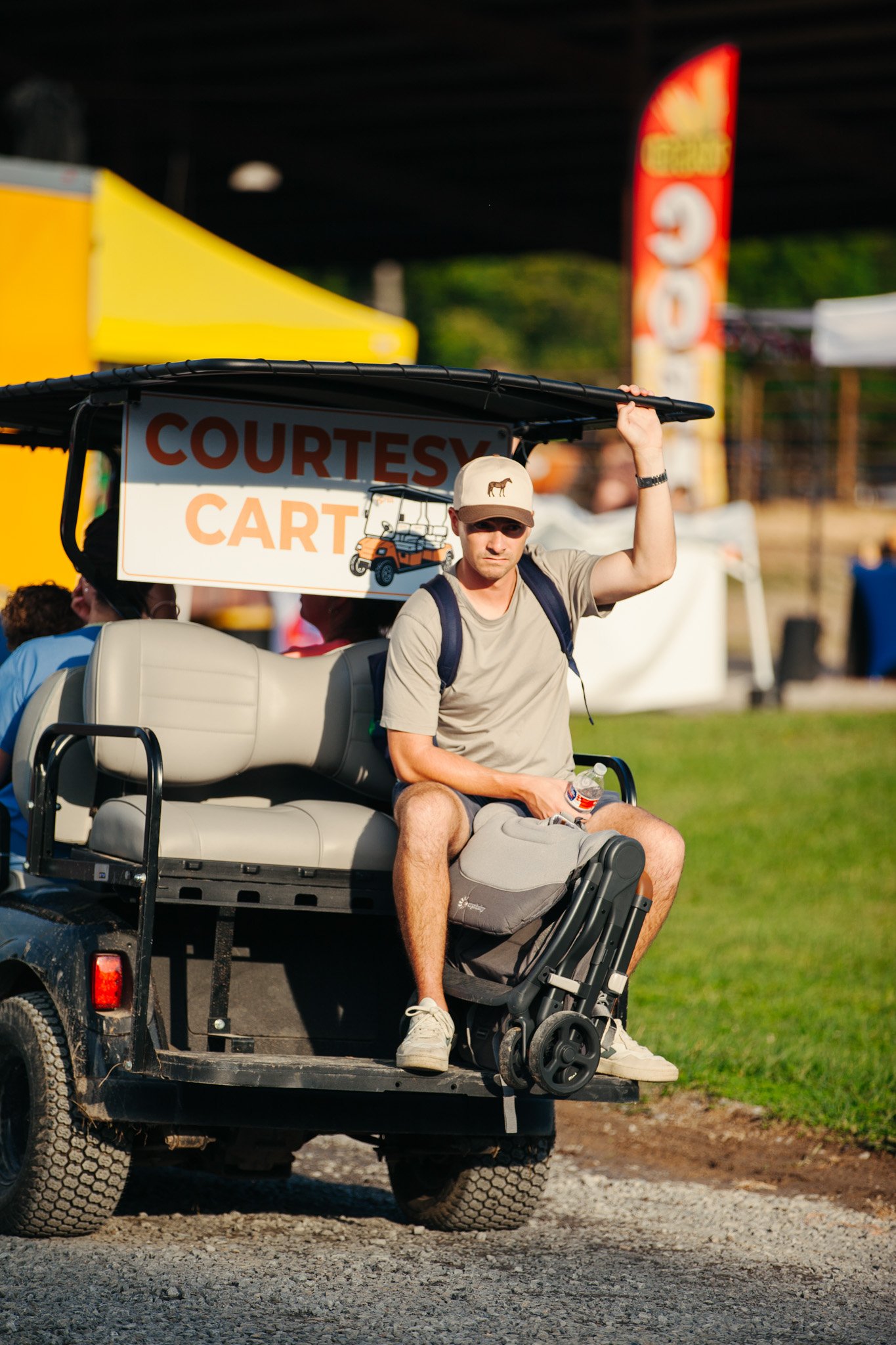 Man sitting on a golf cart wearing a beige cap, khaki shirt, and gray shorts, holding a water bottle, with a 