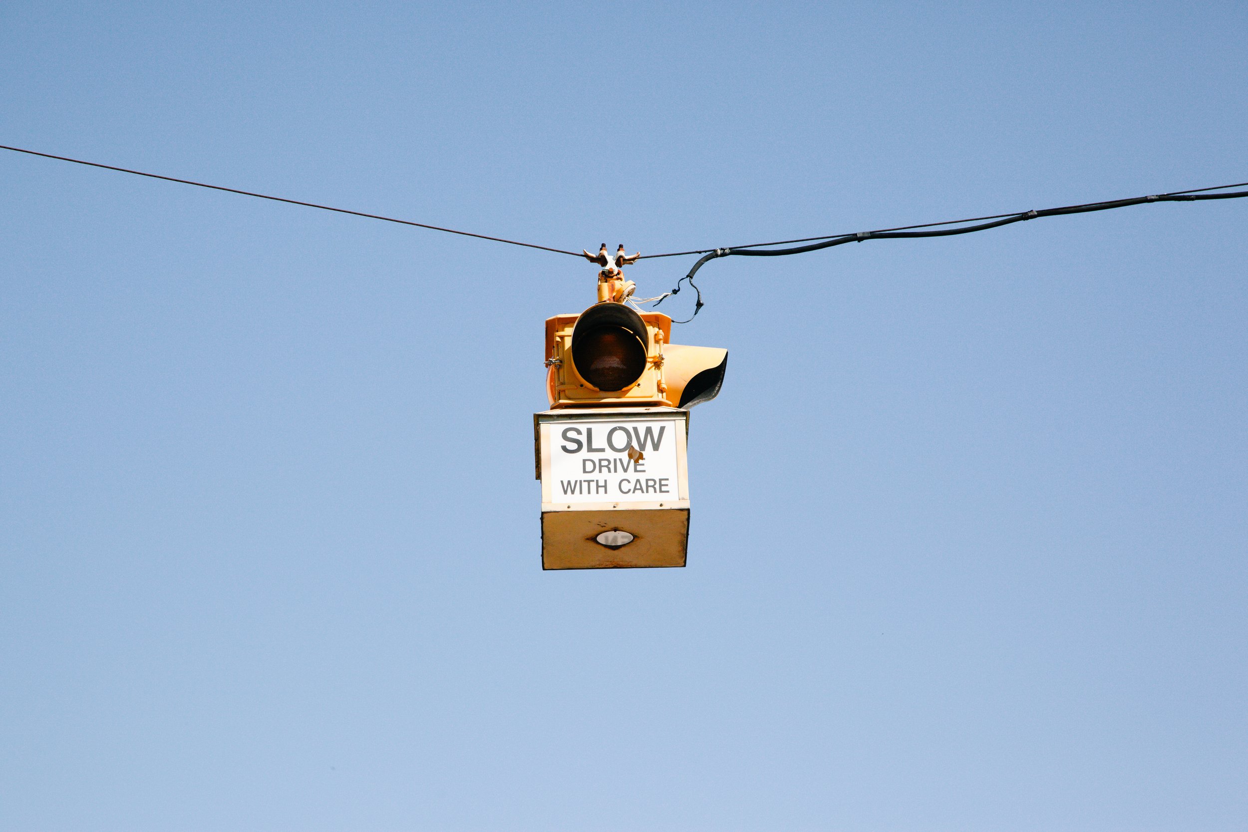 A construction work platform with a traffic signal and a sign that reads 'Slow Drive With Care,' suspended on cables against a clear blue sky.