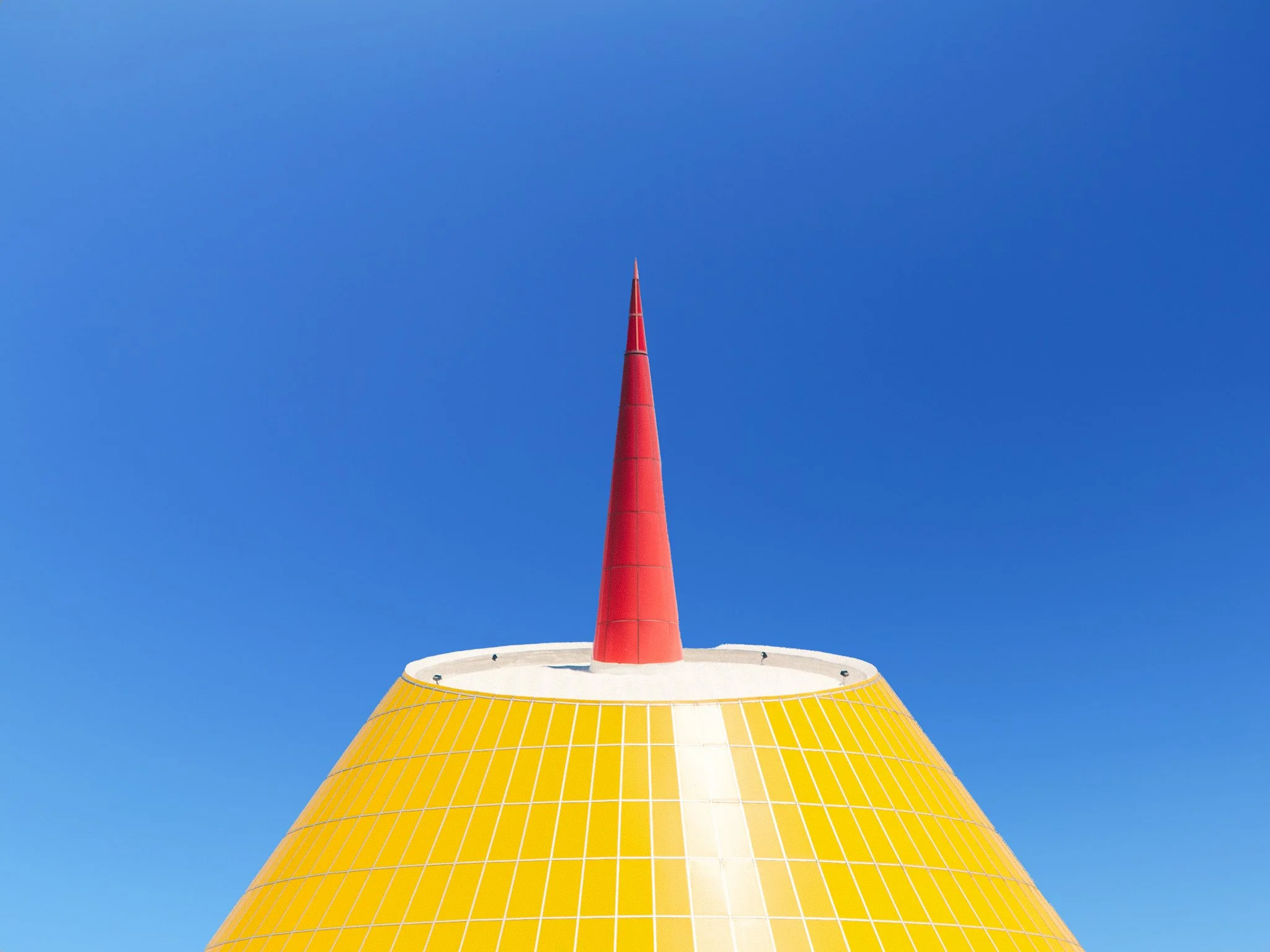 A colorful, modern structure featuring a yellow and white geometric base and a tall red spire against a clear blue sky.