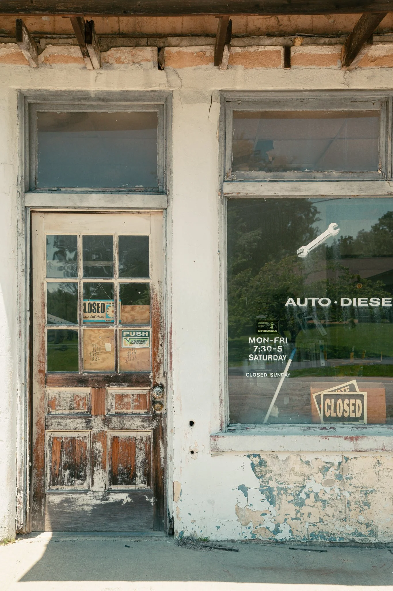 Old, weathered storefront with a wood and glass door and a large window, both showing signs of peeling paint and decay, with 'closed' signs displayed, and a damaged brick and plaster exterior.