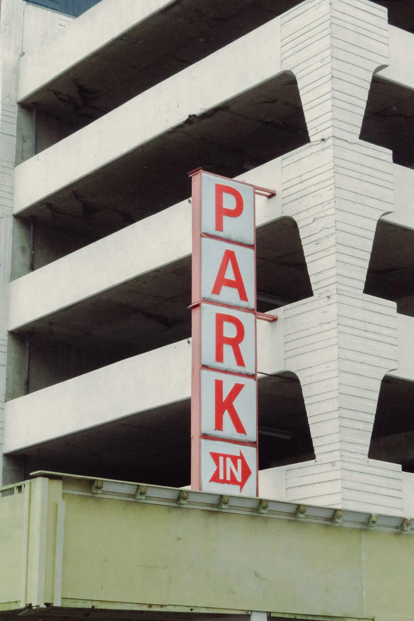Multi-story parking garage with a vertical red and white sign reading 'PARK IN' with an arrow pointing to the right.
