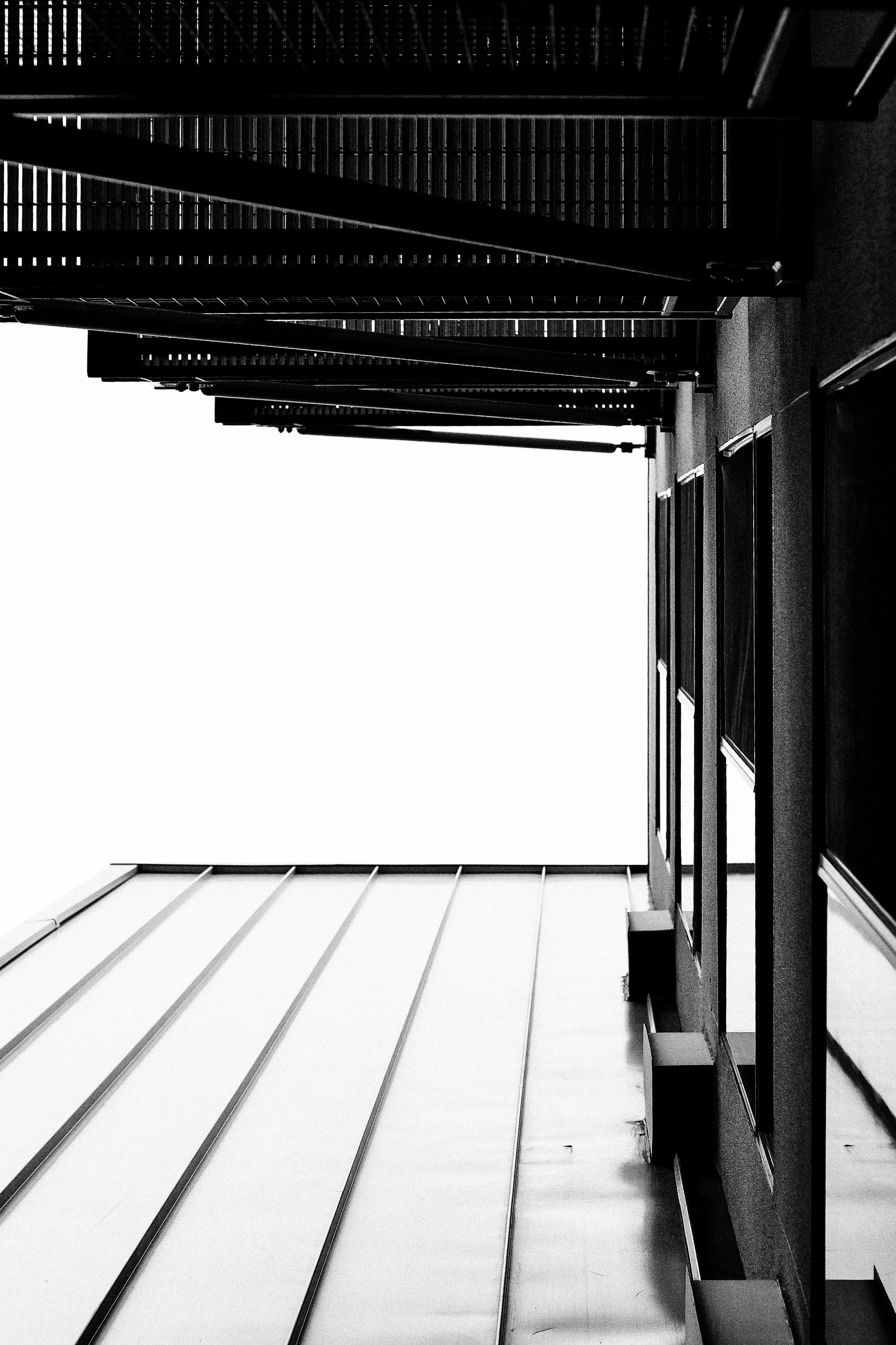 Black and white photo of an indoor tiled space with a railing on the right side and steps below, leading upward to an illuminated space with a grid-like ceiling.