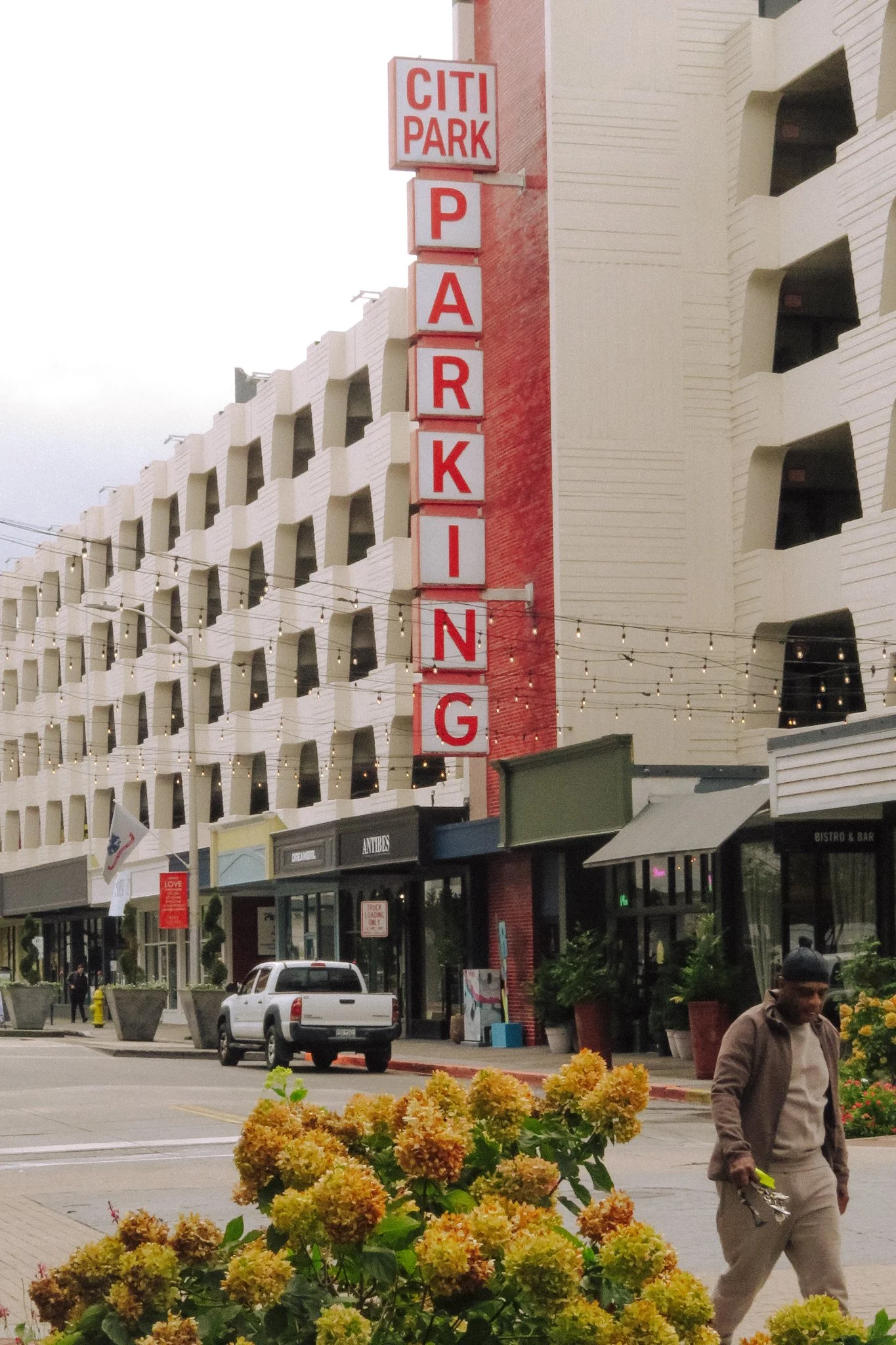A city street scene with a tall vertical sign displaying 'CITI PARKING' in red letters on a white background, and a multi-story white building with balconies in the background. There are storefronts at street level, string lights hanging overhead, an