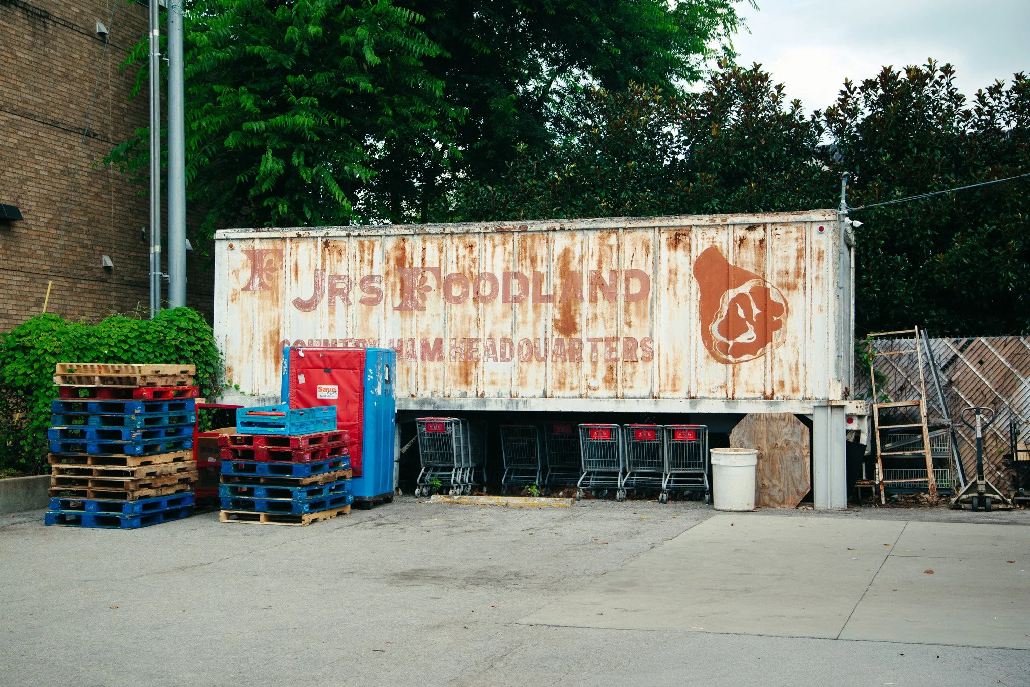A rusty food pantry container with the words 'JRS FOODLAND' faintly visible, surrounded by stacked empty plastic pallets, shopping carts, and a trash bin, located outdoors on a concrete surface with green trees and a brick building in the background.