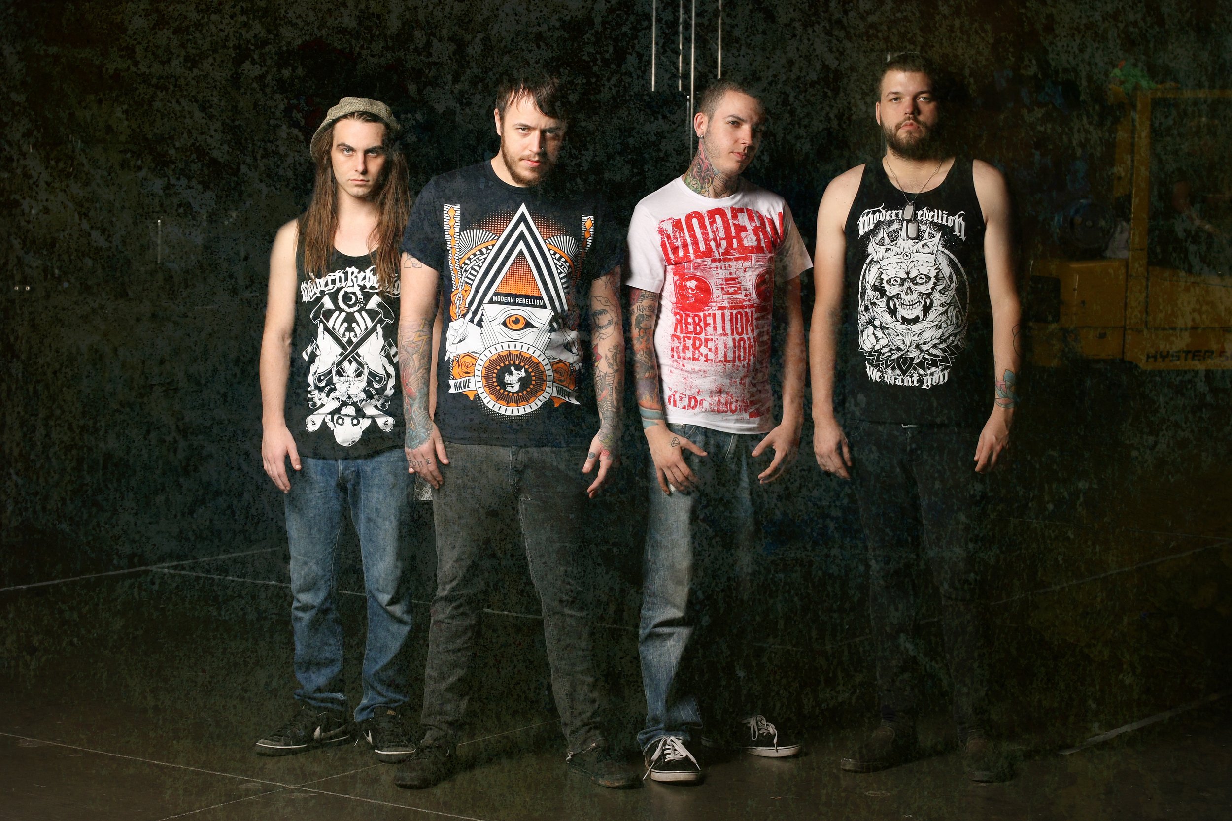 Four young men with tattoos standing against a dark background, wearing graphic tank tops and jeans.