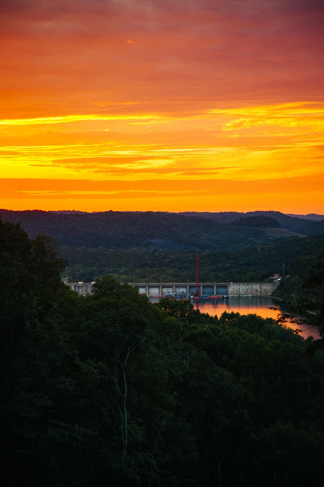 A sunset over hills and a river, with an industrial dam and crane visible in the distance, surrounded by lush green trees.