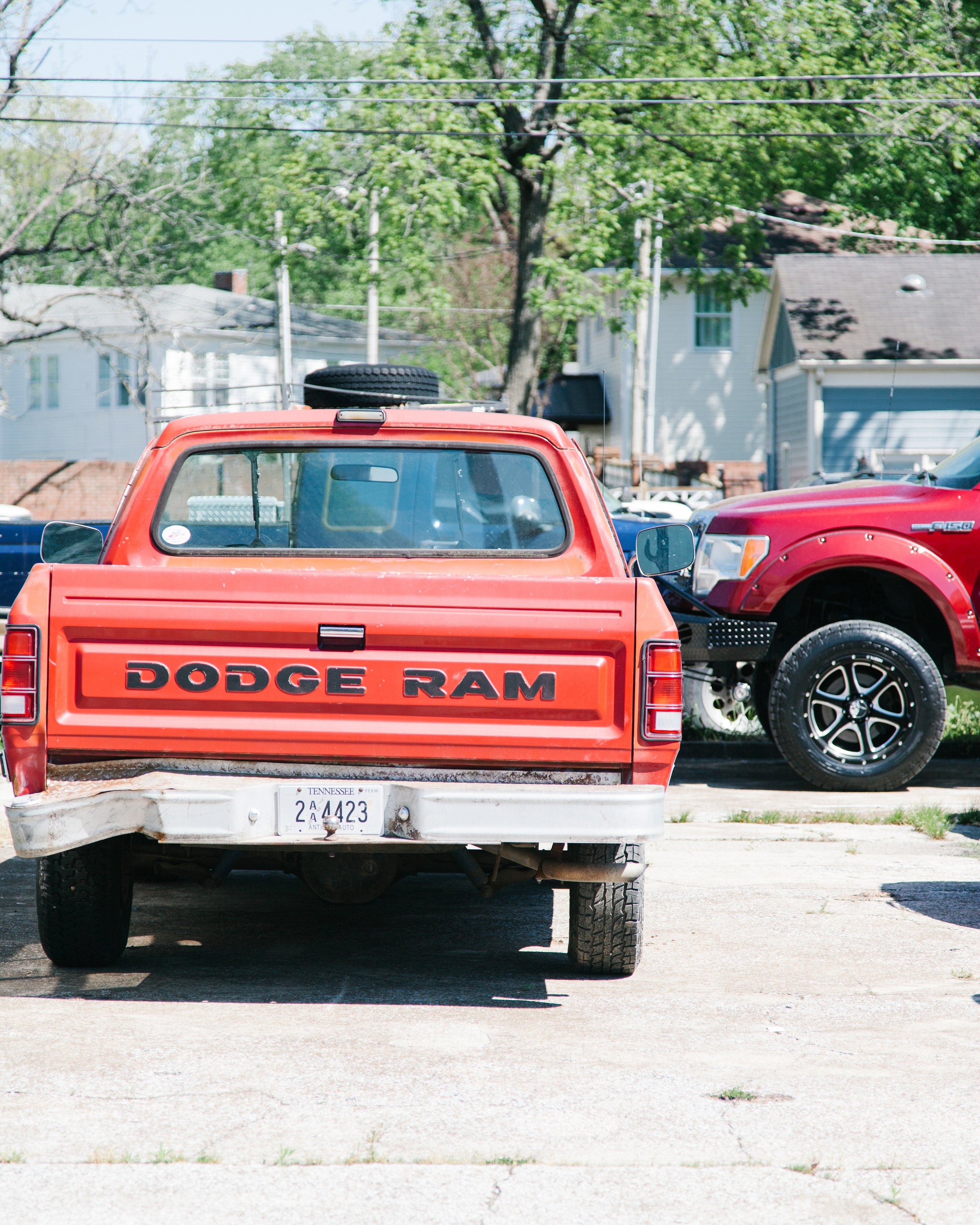 Rear view of a red Dodge Ram pickup truck parked on a concrete lot with another red pickup truck to the right and trees and houses in the background.