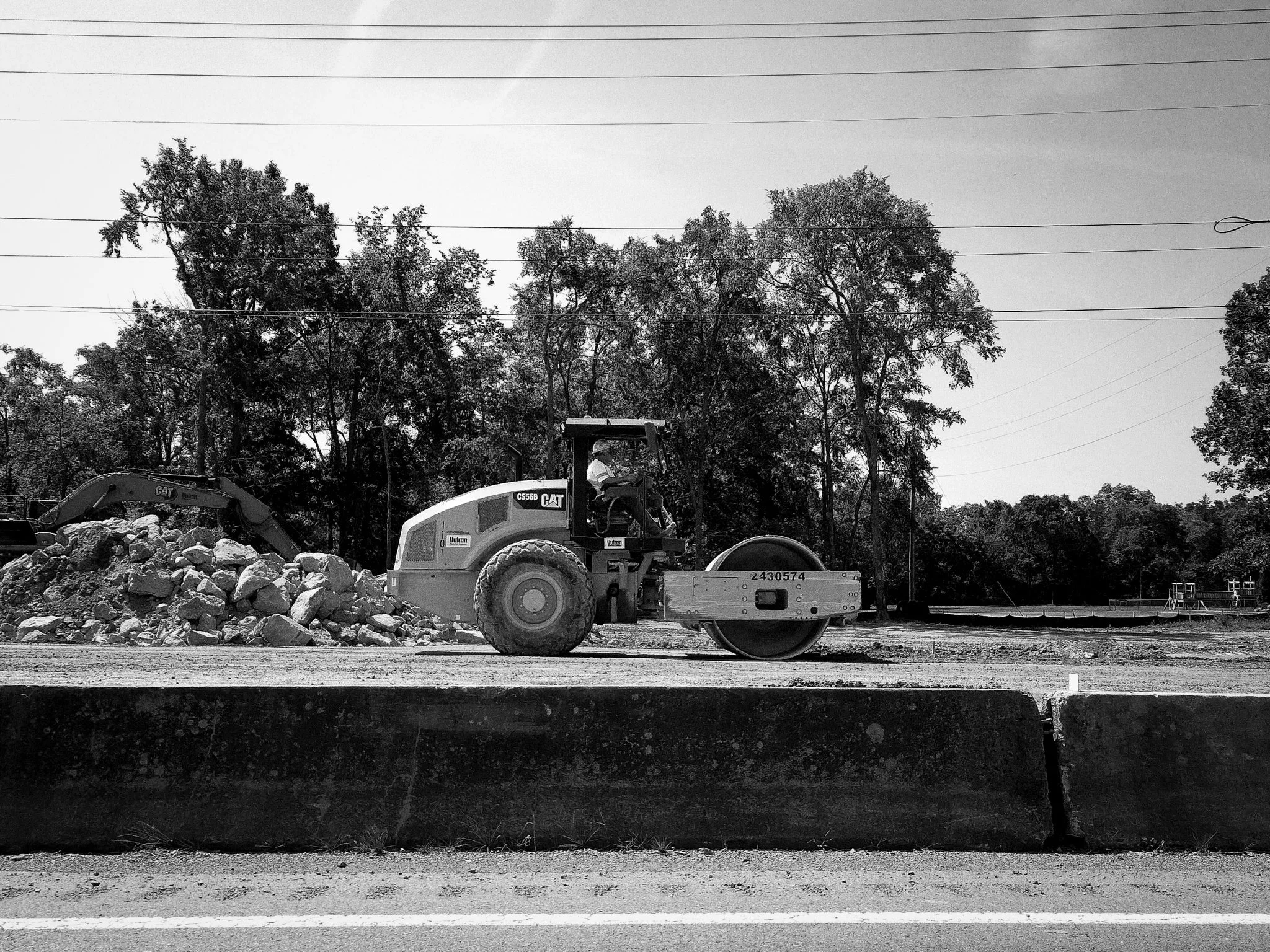 Black and white photo of construction site with a Caterpillar compact excavator moving rocks, trees in the background, and electrical wires overhead.