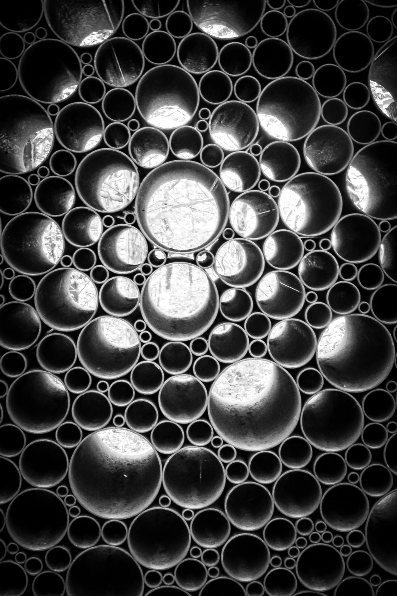 A close-up of various sizes of circular pipes arranged together, with some reflecting light and showing a glimpse of trees or sky through the openings.
