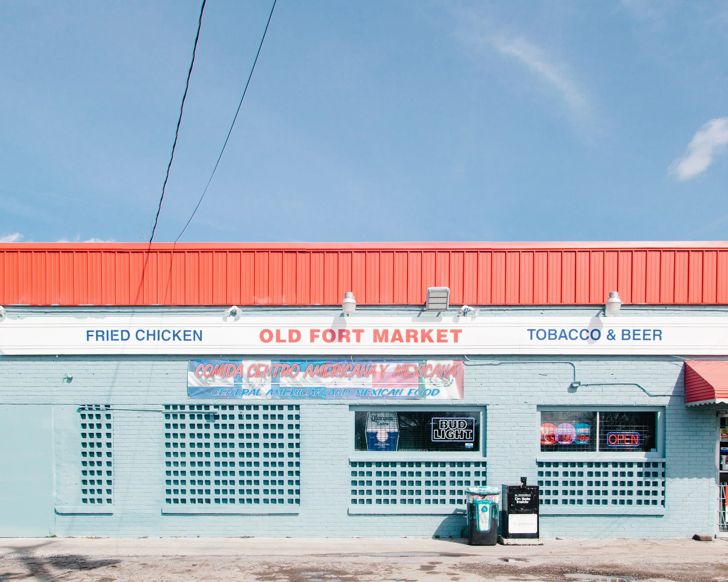 The storefront of Old Fort Market, a convenience store with a neon sign advertising Bud Light, and signs for fried chicken, tobacco, and beer. The building features a red roof and a white brick exterior with a few small advertisements and an open sig