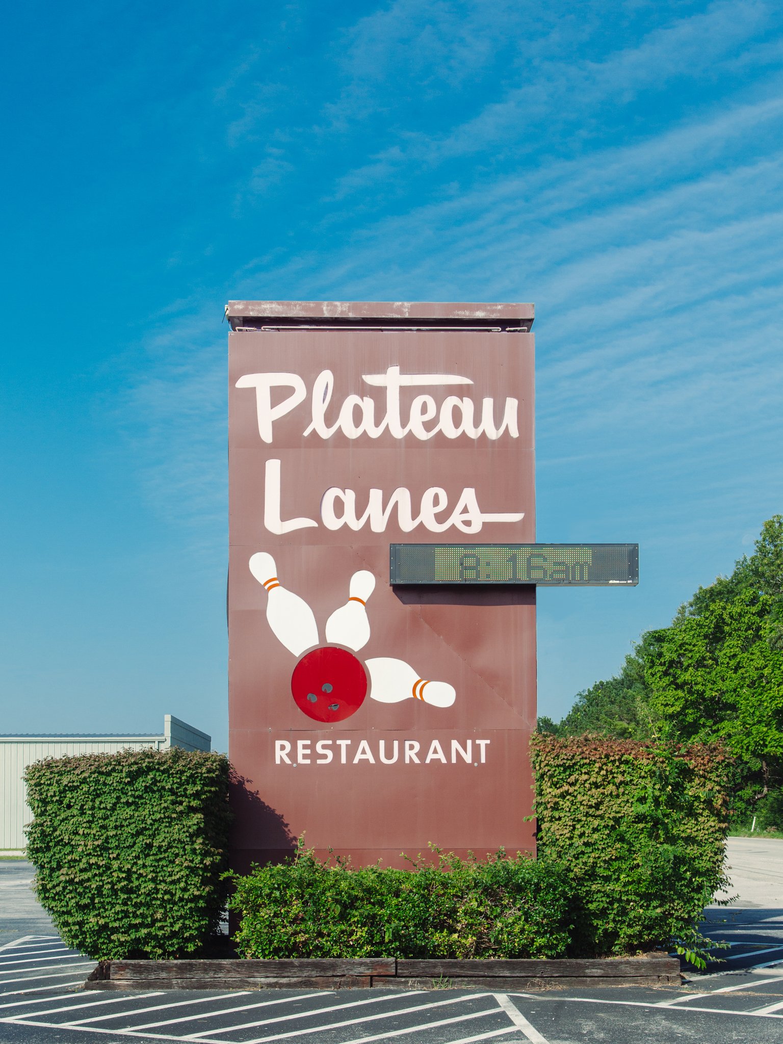 Sign for Plateau Lanes Restaurant with a bowling ball and pins illustration, set against a clear blue sky and surrounded by green bushes.