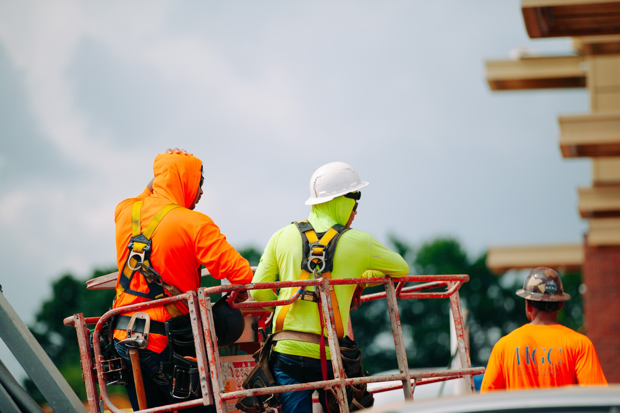 Construction workers wearing safety gear, including helmets and high-visibility clothing, are working on a construction site. They are on an elevated platform.