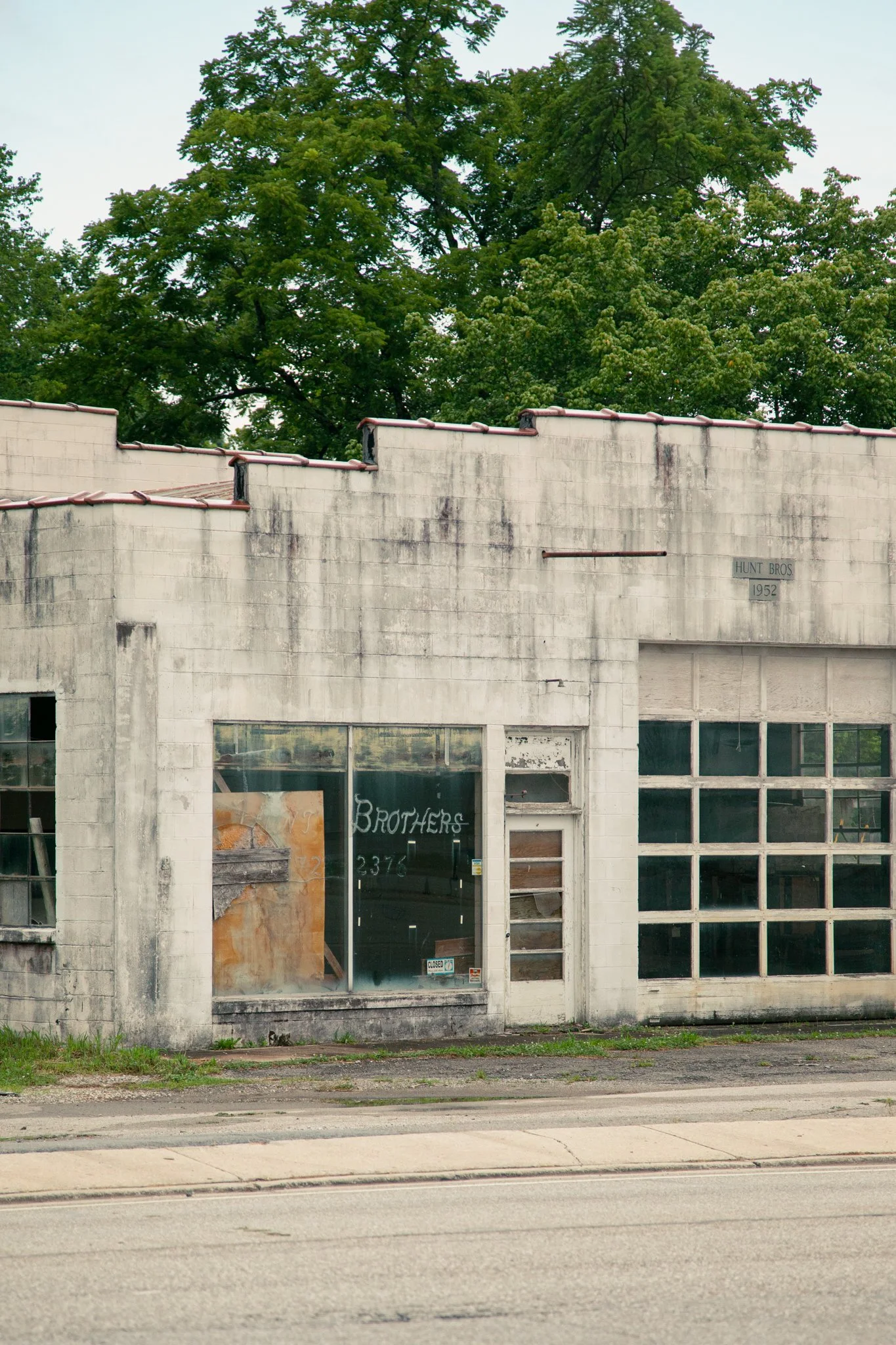 An abandoned storefront with broken windows and faded signage, overgrown grass in front, with large trees in the background.