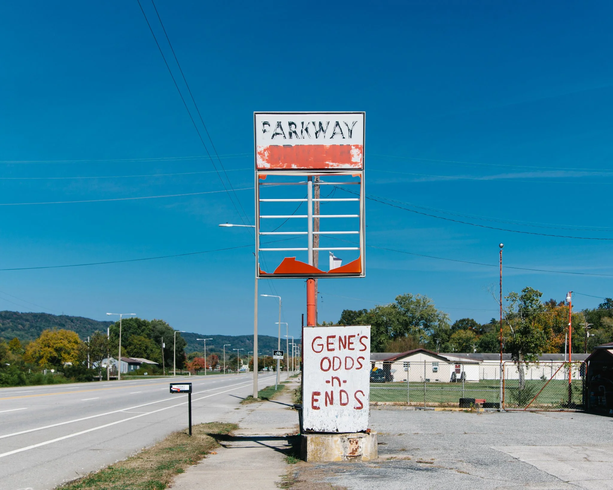 A roadside billboard with a peeling sign that reads 'GENE'S ODDS END' in red letters, with a partly destroyed lower section. There is also a faded larger sign above that says 'PARKWAY,' which is damaged and difficult to read. The scene depicts a rura