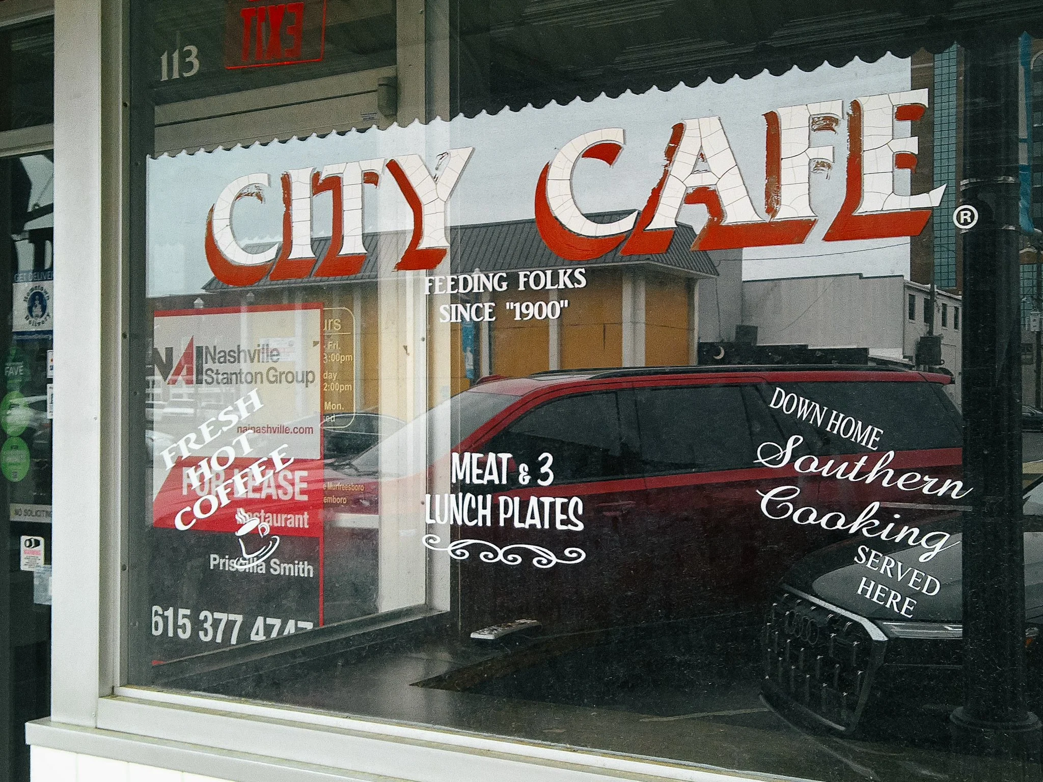 View of a restaurant window displaying the name 'City Cafe' with the tagline 'Feeding Folks Since 1900'. Additional signs mention breakfast, lunch plates, and a phone number. The window has some reflections of the street and parked cars outside.