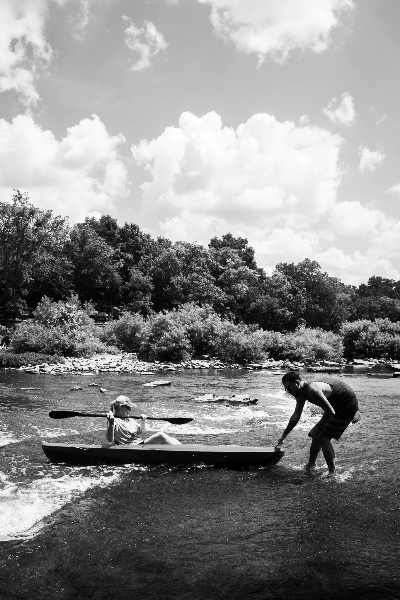 A man and a woman in a small boat on a river, with the man helping the woman out of the boat onto the riverbank, surrounded by trees and a cloudy sky.