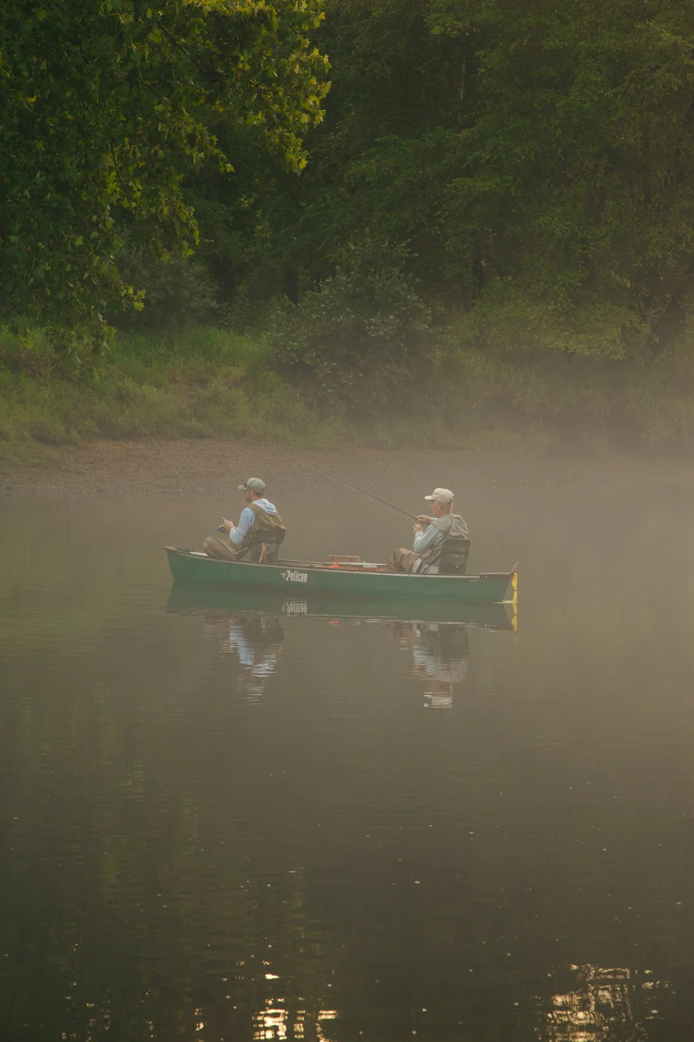 Two men fishing from a small boat on a calm river, surrounded by dense green trees, with a soft fog or mist over the water.
