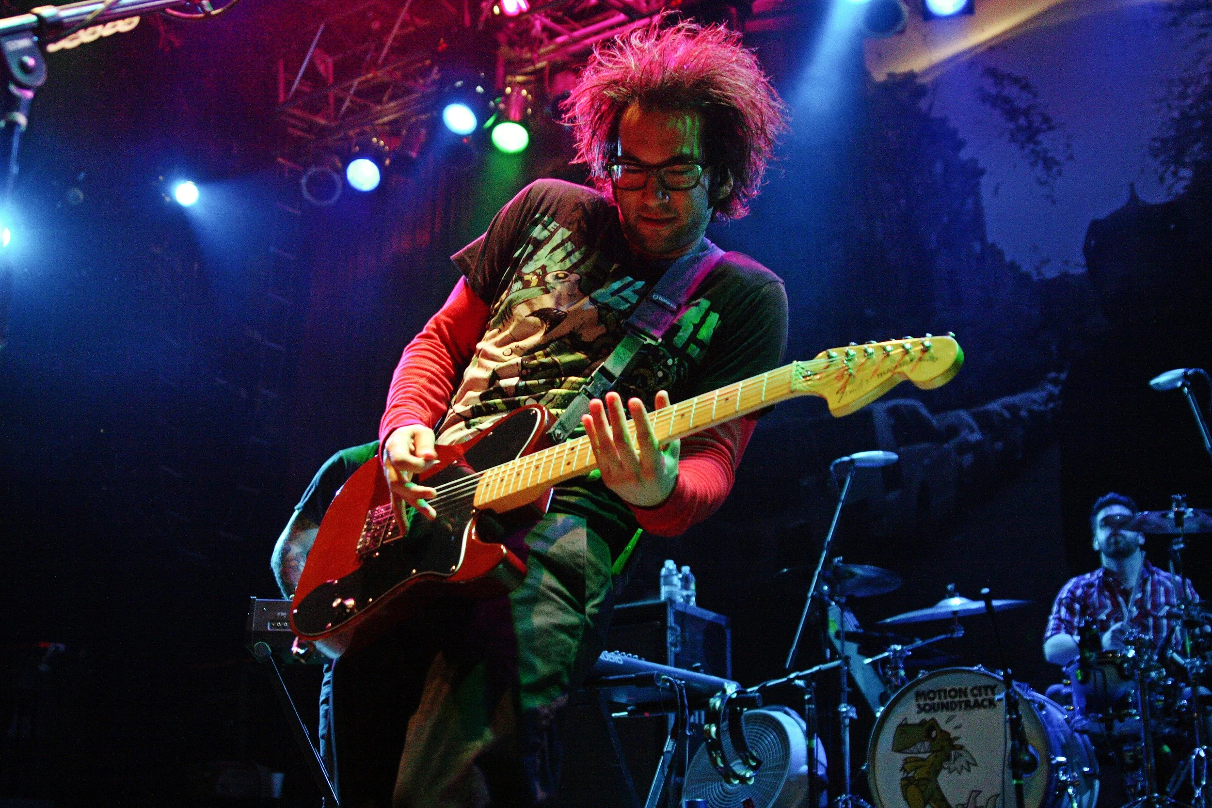 A musician playing a red electric guitar on stage under colorful lights. The musician has messy hair, glasses, and a graphic t-shirt. A drum set is visible in the background with the text 'Motion City Soundtrack.'