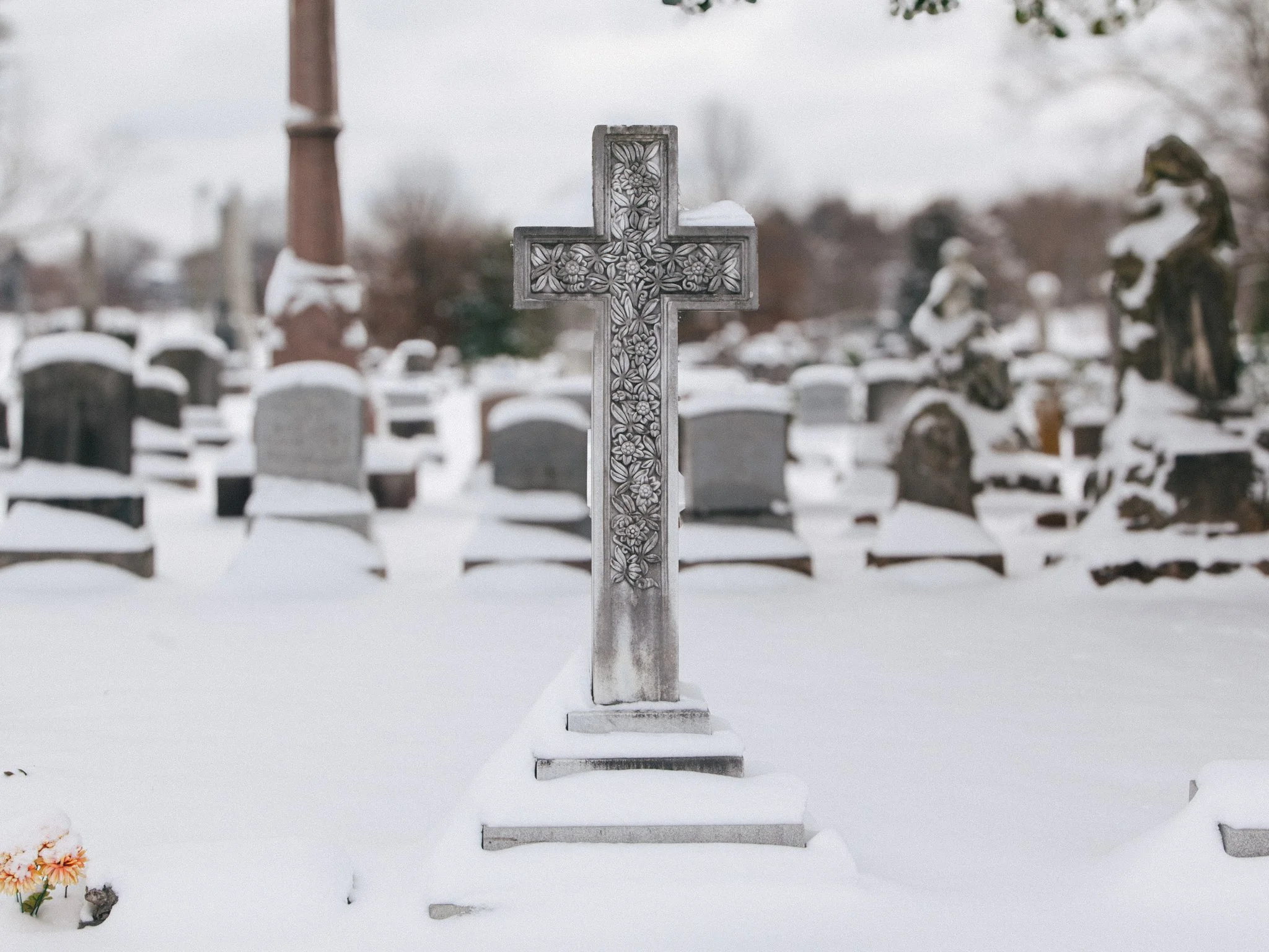 A snow-covered cemetery with a decorative metal cross in the center, surrounded by tombstones and snow, with blurred trees and structures in the background.