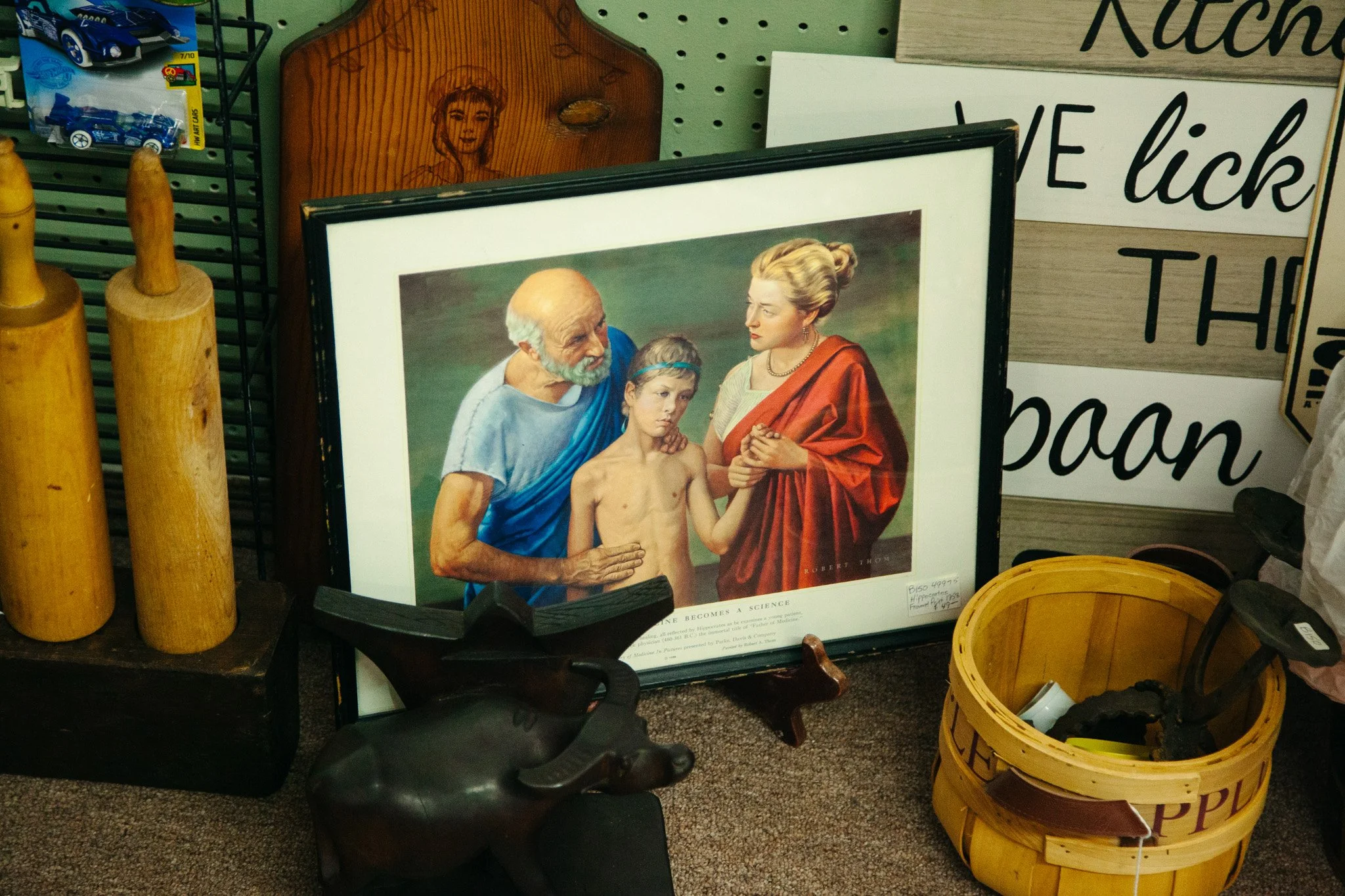 A framed poster of Leonardo da Vinci's 'The Vitruvian Man' with people in the background; surrounding items include yellow wooden rolling pins, a black sculpture of a dog's head, and a yellow basket. There are also signs with text on the wall that ar