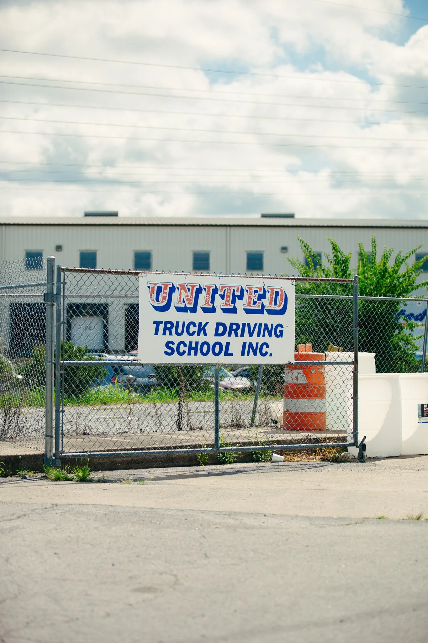 A chain-link fence with a sign that reads 'United Truck Driving School Inc.' in front of a large industrial building with a metal roof, some green trees, and a partly cloudy sky.
