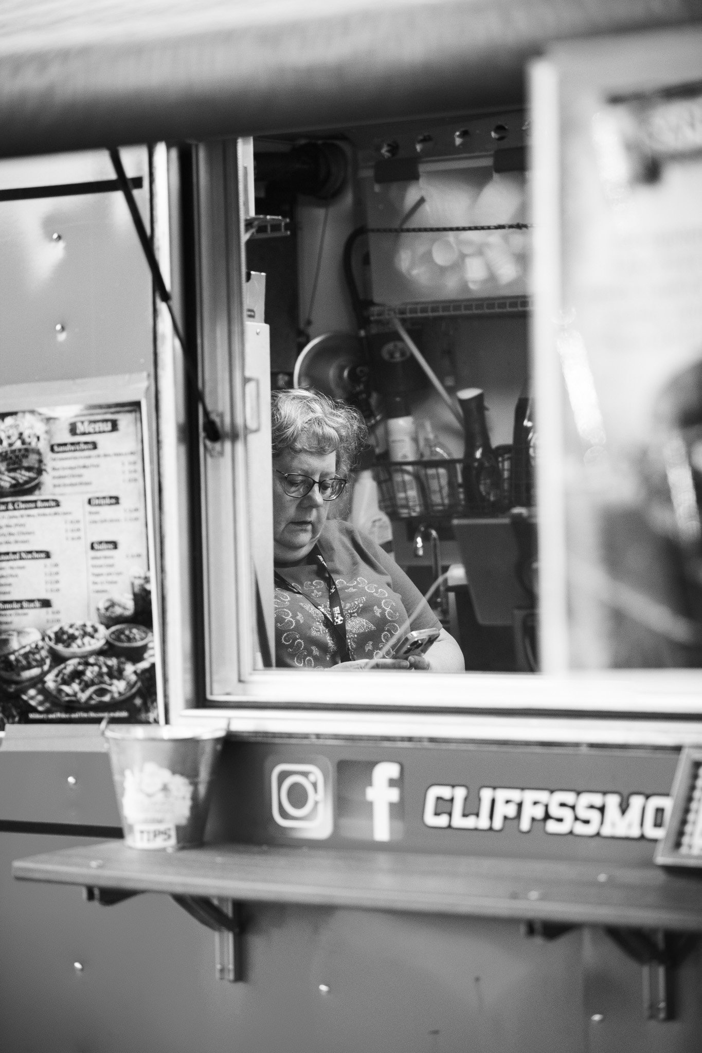 A woman with glasses and curly hair working at a food kiosk, looking at her phone, with a menu and social media stickers on the counter.
