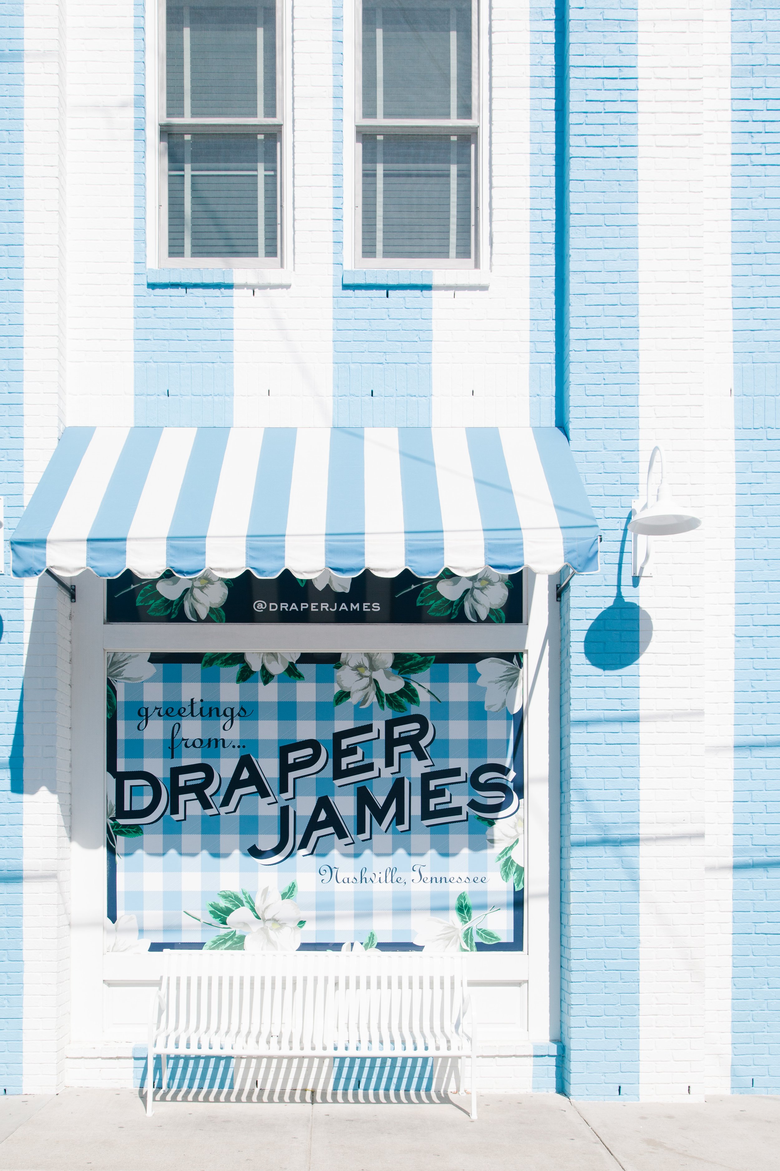 Storefront with a blue and white striped awning and a sign that reads 'Greetings from ... Paper Jams.' The building has white brick walls with light blue accents, and there are two windows above the awning.