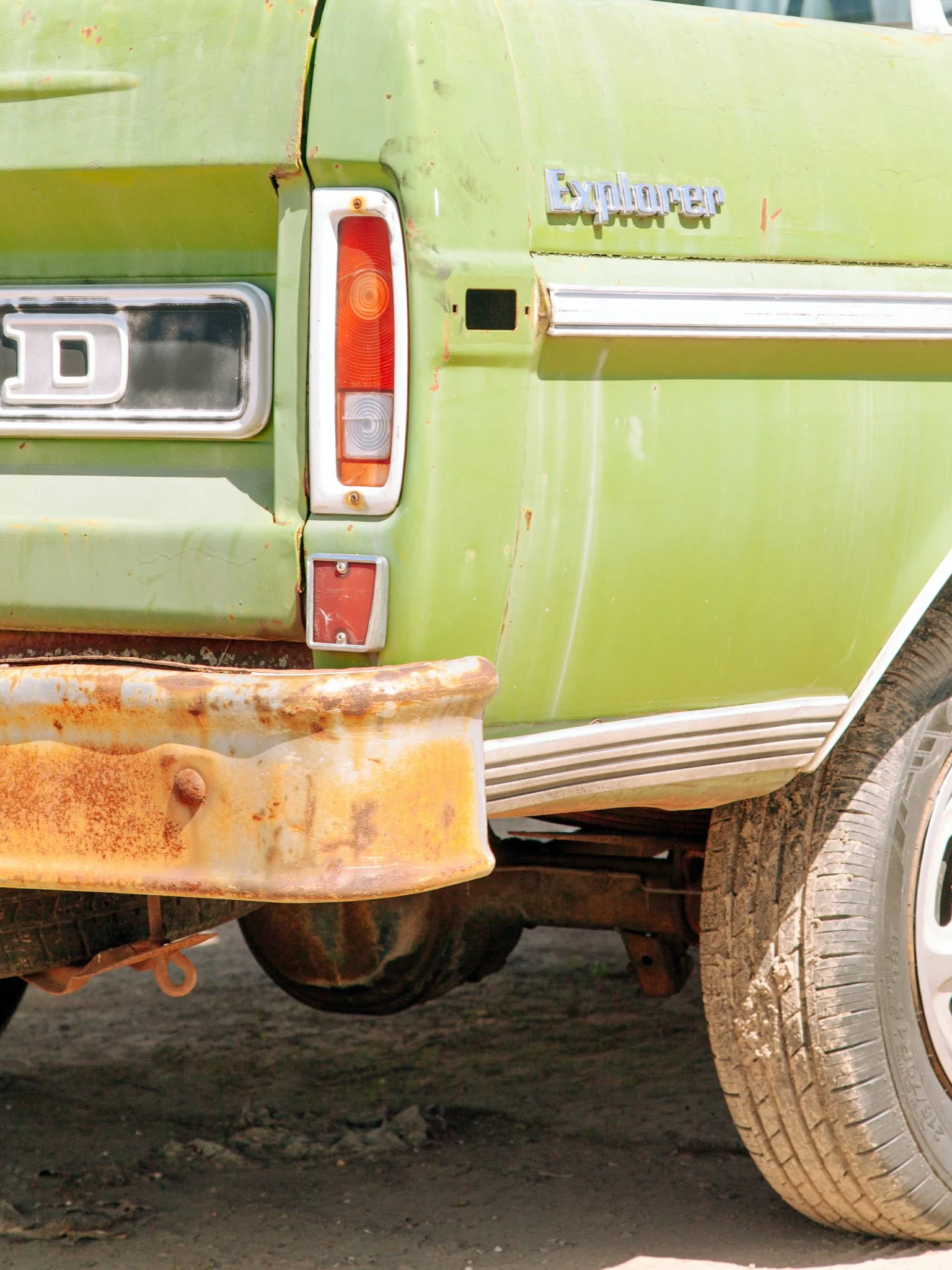 Close-up of the back of an old, weathered green Ford Explorer SUV with rust and dirt, showing tail lights, bumper, and part of the rear wheel.