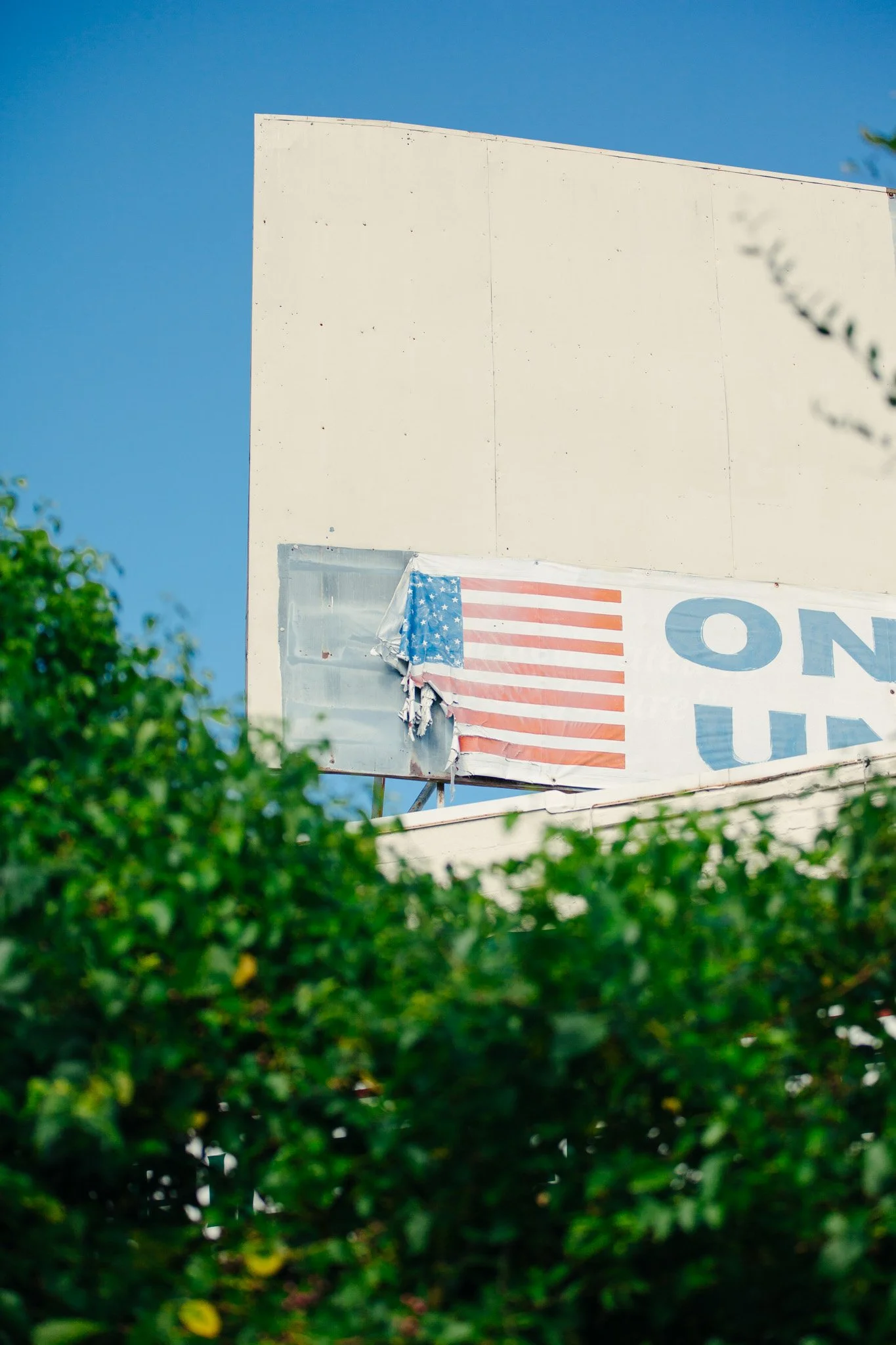 Part of a billboard with an American flag design and blue text, partially obscured by green leaves in the foreground, against a bright blue sky.