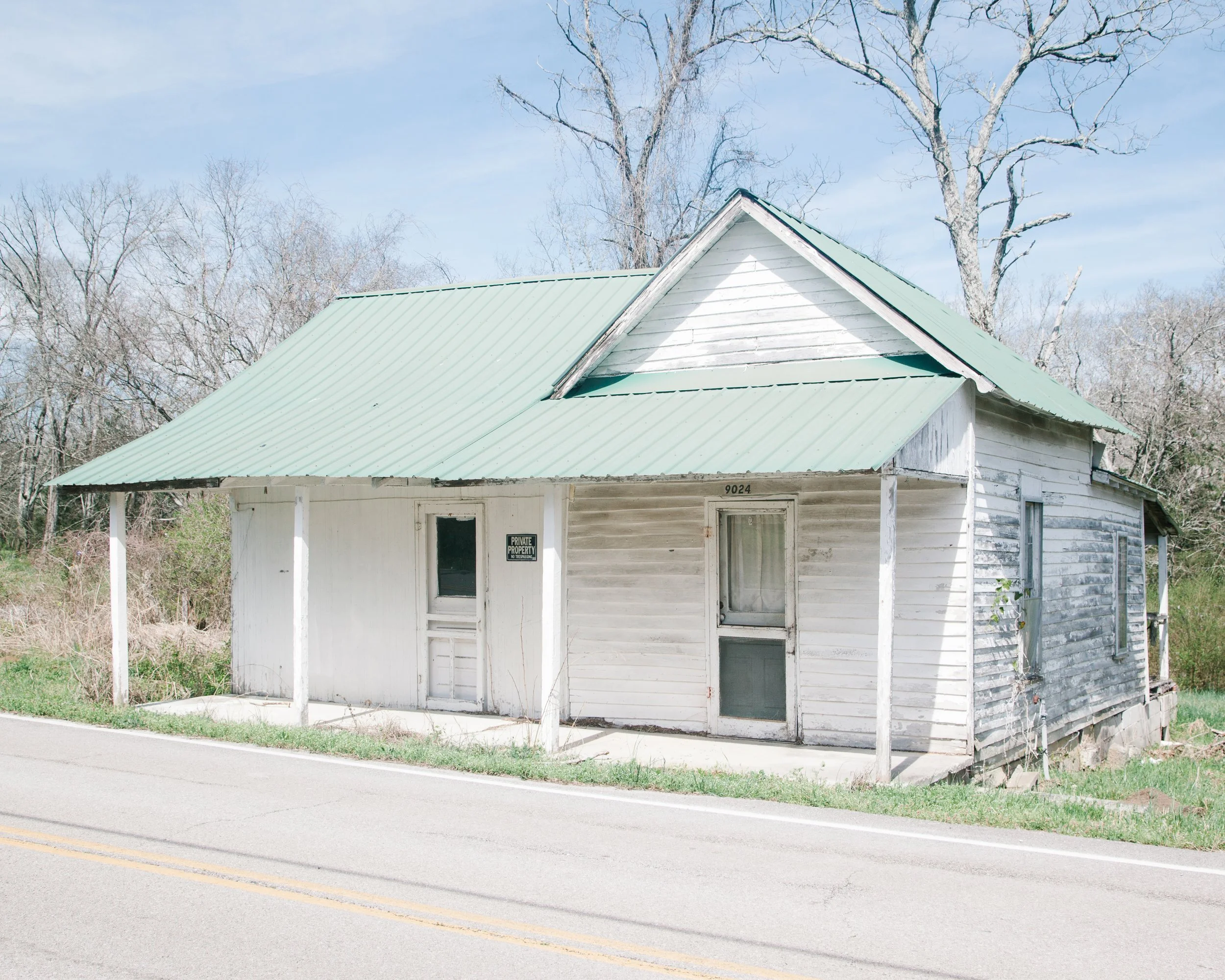 A small, weathered, white house with a green metal roof, located next to a street with double yellow lines. The house has a front porch with a sign that reads 'Private Property' and some windows. Behind the house, there are leafless trees and a partl