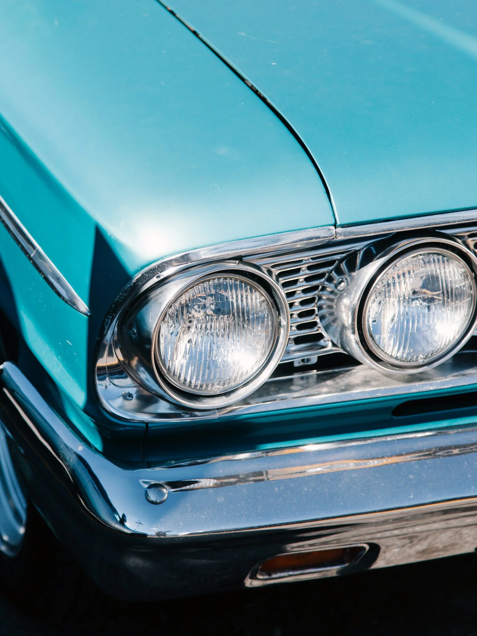 Close-up of a vintage teal car's front end, showing dual round headlights, chrome detailing, and part of the hood.