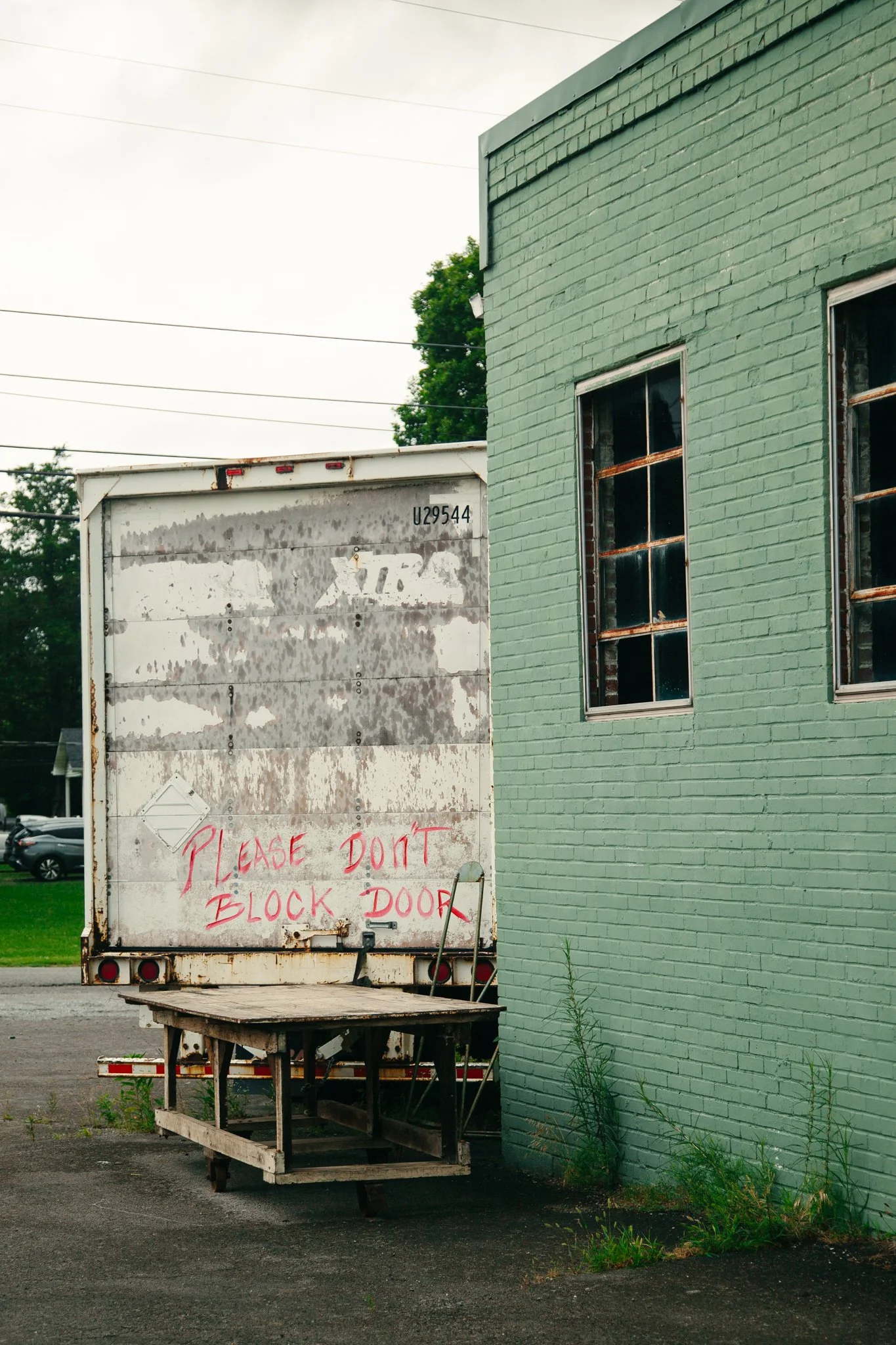 A white cargo truck with a weathered, rusted door is parked next to a green brick building. A handwritten sign in red says, 'Please don't block door' on the back of the truck. There is a flatbed trailer in front of the truck with some overgrown weeds
