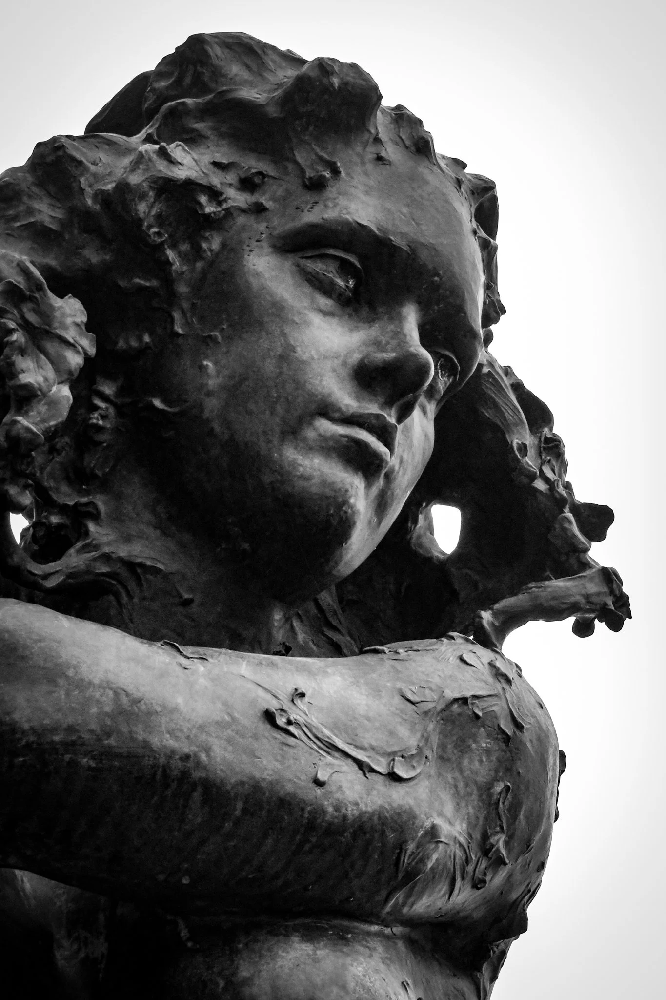 A close-up view of a bronze statue of a woman's face and upper body, with detailed hair and facial features, set against a plain white background.