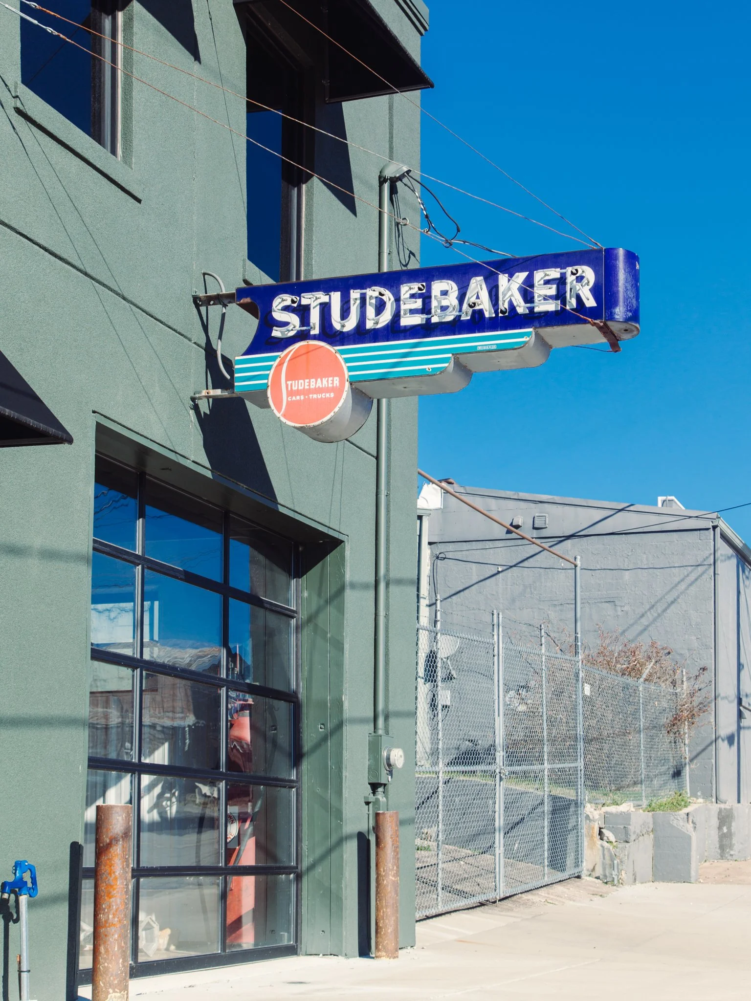 The photo shows the exterior of a building with a green wall and large glass windows. There is a blue and white sign hanging from the building that reads 'STUDEBAKER' with an orange circle below that says 'TUDEBAKER CARS-TRUCKS'. The building has a b